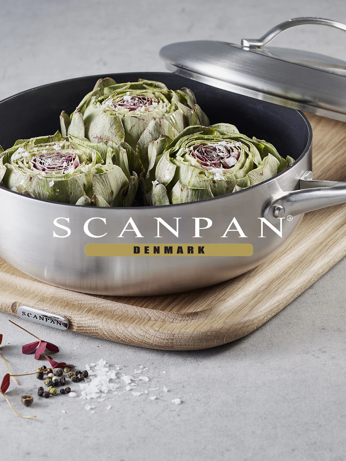 Kolme halkaistua artisokkaa suolan kera harjatussa teräskattilassa puulaudalla. Hajallaan pippuria ja suolahiutaleita lähellä.