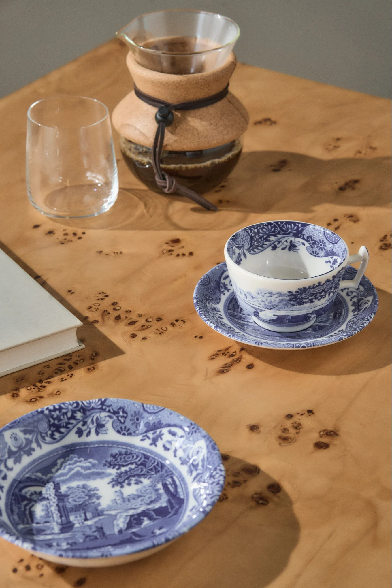 Pour-over-kahvinkeitin, tyhjä lasi, sinivalkokuvioinen teekuppi lautasella ja yhteensopiva lautanen vaalealla puupöydällä.