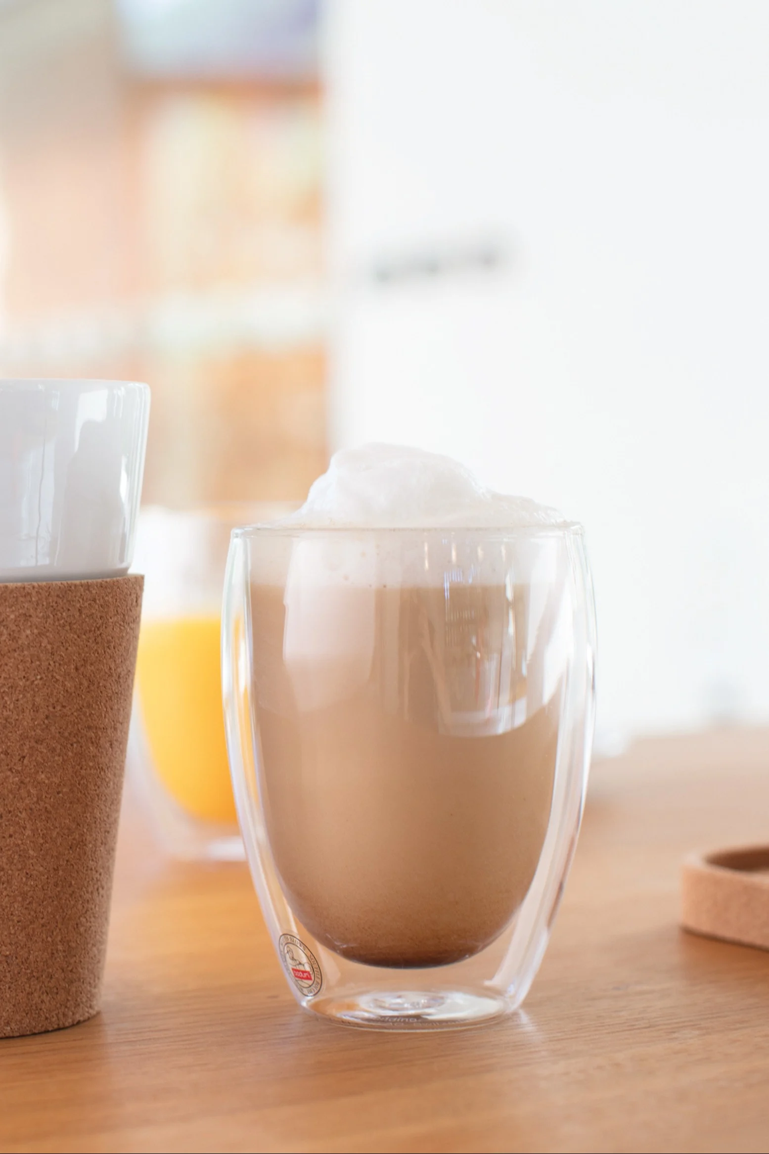 Vaahdotettu latte kaksiseinäisessä kirkkaassa lasimukissa puupöydällä, toinen muki korkkihylsyllä vasemmalla ja appelsiinijuoma taustalla.