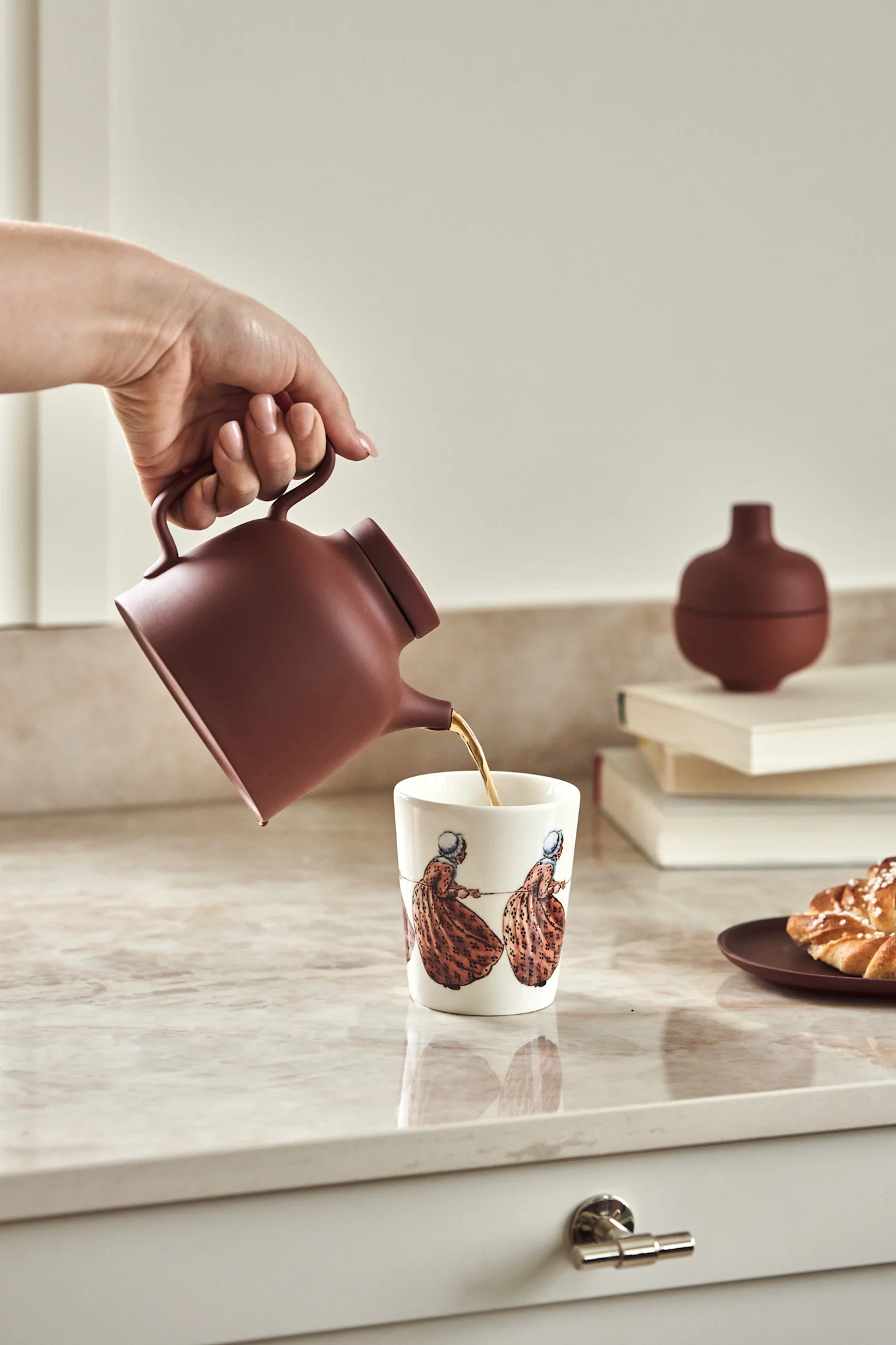 Muki, jossa on ruotsalaisen kirjailijan Elsa Beskowin Aunt Brown -motiivi keittiön tiskipöydällä yhdessä terrakotanpunaisen Sand-kannun kanssa.