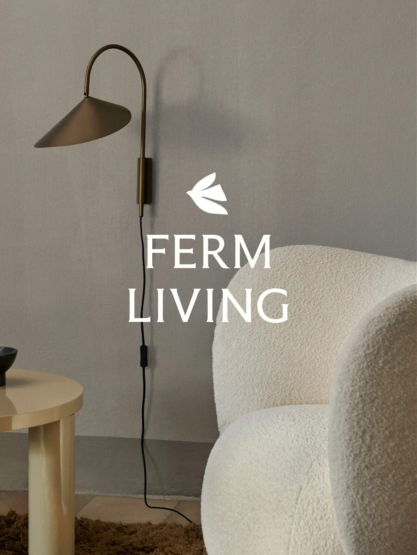 Ferm Living -logo messinkisen seinävalaisimen, kermanvärisen bouclé-nojatuolin ja vaalean puisen sivupöydän päällä minimalistisessa sisustuksessa.