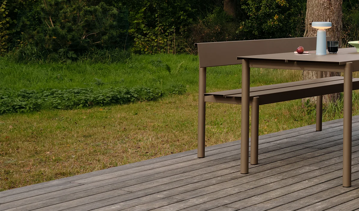 Moderni taupenvärinen ulkoruokapöytä ja penkki puukannella, pöydällä kannettava lamppu ja omenia.