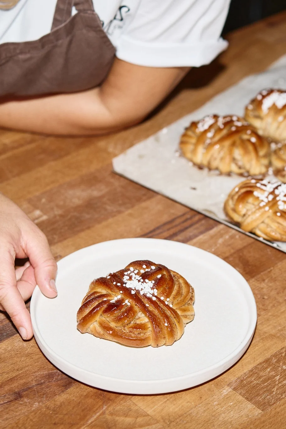 Käsi asettaa sokeroidun, punotun korvapuustin lautaselle; lisää pullia tarjottimella.