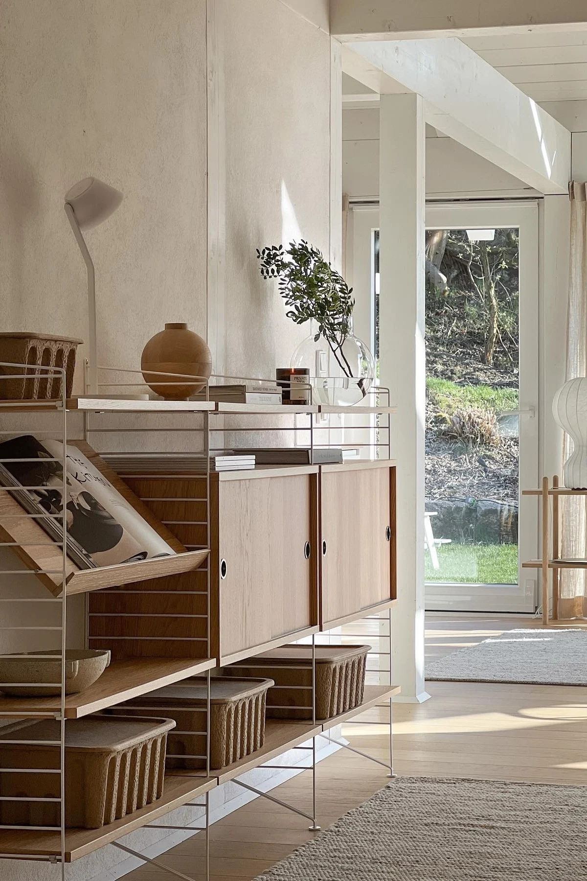 Valoisa, moderni huone, jossa on vaaleasta puusta ja valkoisista kehyksistä koostuva String-hylly, täynnä maljakkoa, kirjoja, kasveja ja koreja, ja näkymä puutarhaan. Hylly sopii täydellisesti seinän koristeluun.