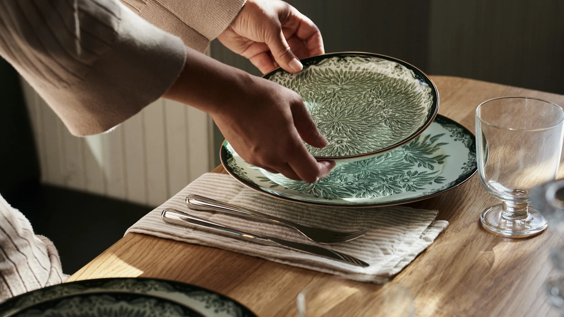 Kädet asettavat vihreä-valkokuvioisen lautasen puupöydälle aterimien ja lasin viereen.