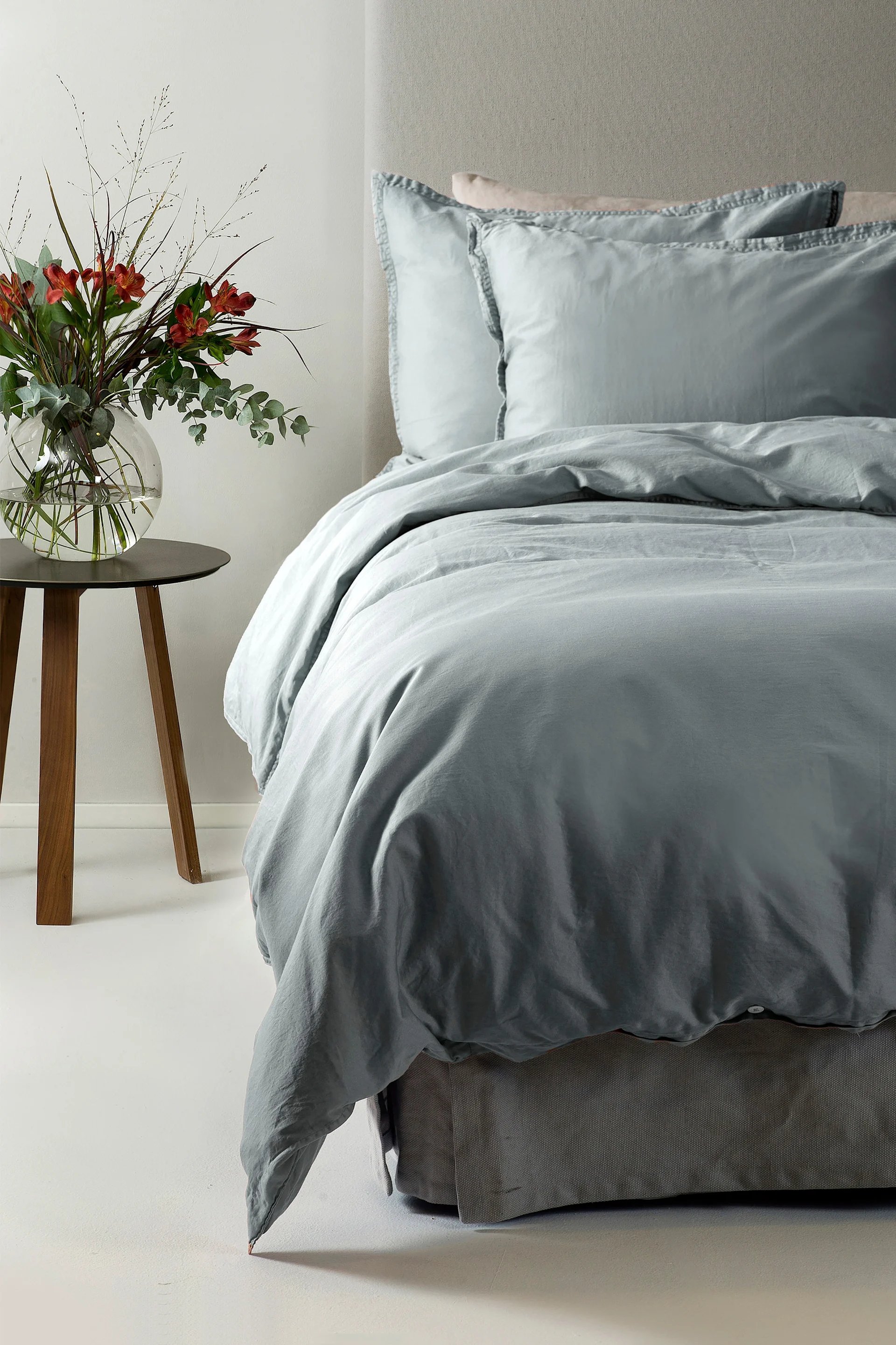 Sänky, jossa on vaaleansiniset vuodevaatteet ja kaksi tyynyä, sekä vieressä puinen pöytä, jossa on maljakko punaisine kukkineen.