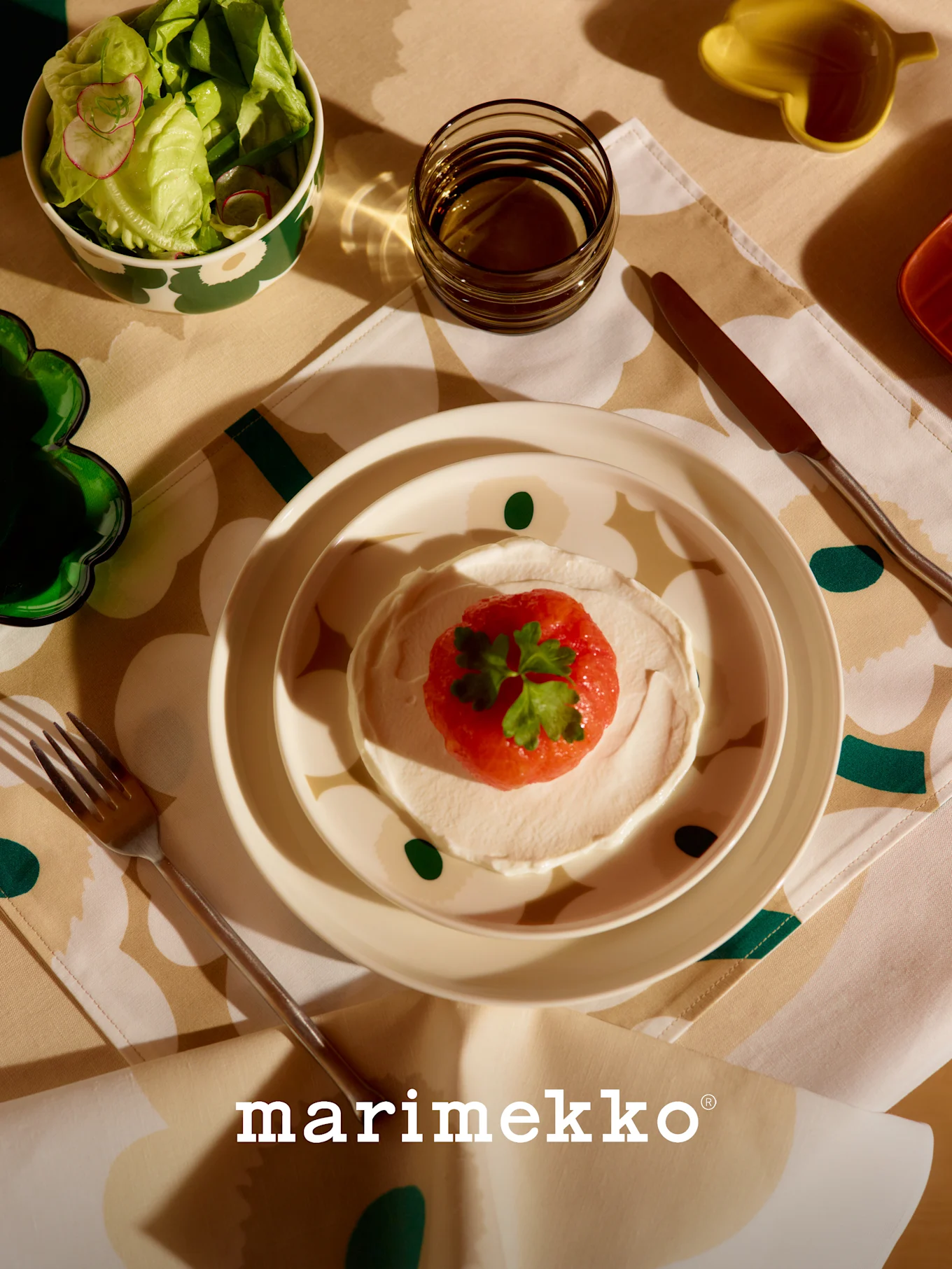 Marimekko-pöytäkattaus, jossa kuviollinen salaattikulho, punainen hedelmä- ja kermaruoka, lasi ja aterimet kukkakuosisella pöytäliinalla.