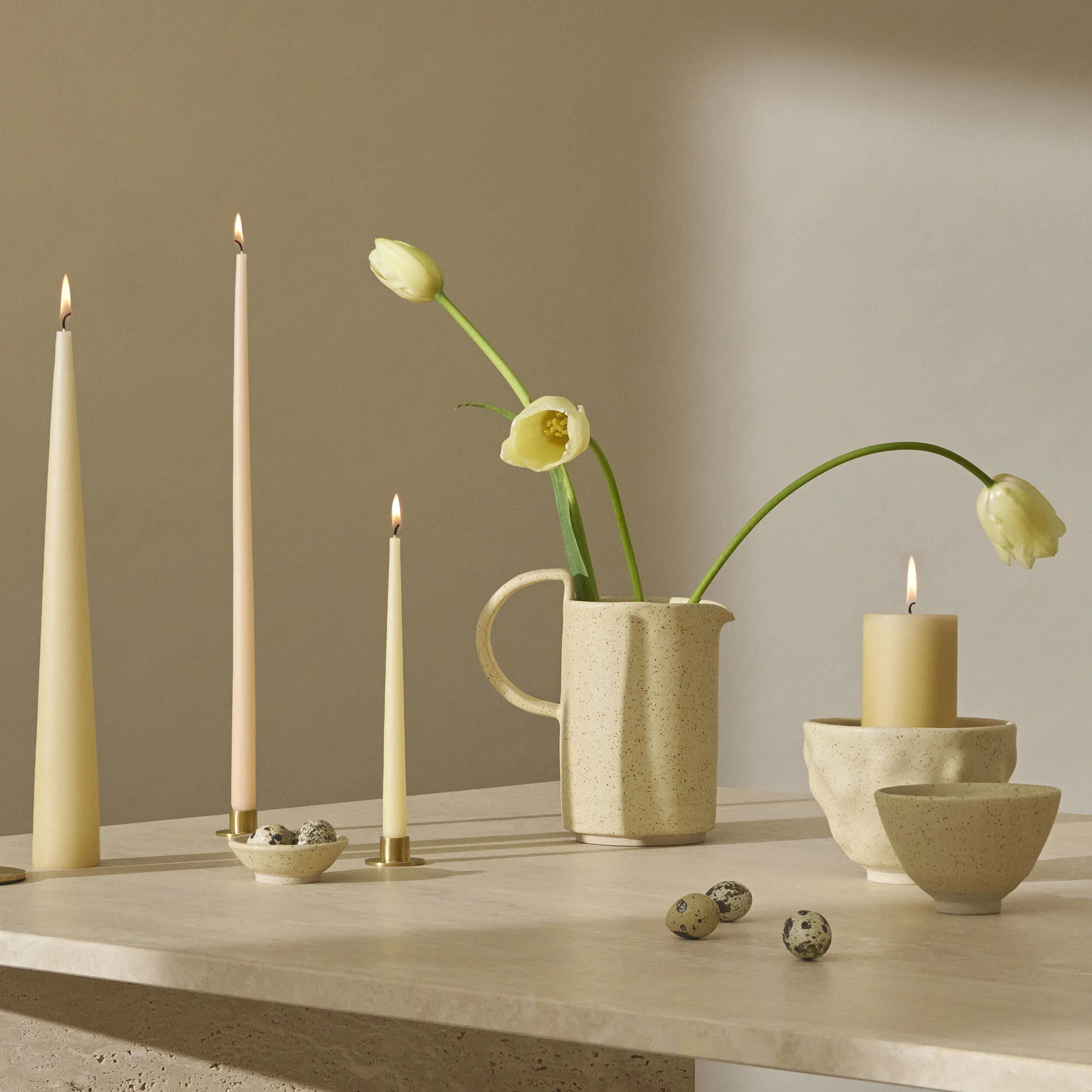 Asetelma sytytettyjen kynttilöiden, vaaleankeltaisten tulppaanien kanssa pilkullisessa kannussa ja viiriäisenmunia tekstuuripinnalla.