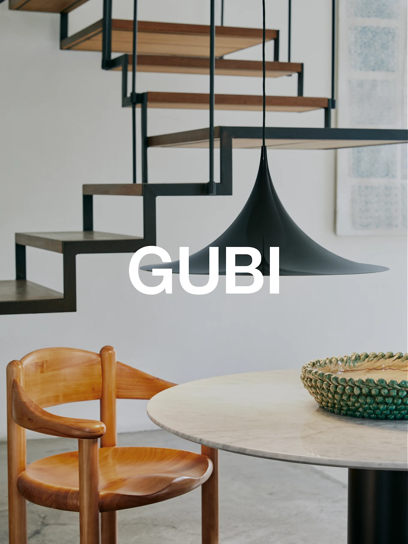 GUBI-logo modernissa sisustuksessa puutuolilla, marmoripöydällä, vihreällä kulholla, mustalla riippuvalaisimella ja moderneilla portailla.