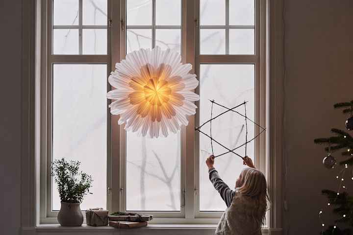 Watt & Veken Snöblomma-joulutähti ikkunassa, jossa lapsi pitelee tikuista tehtyä tähteä.