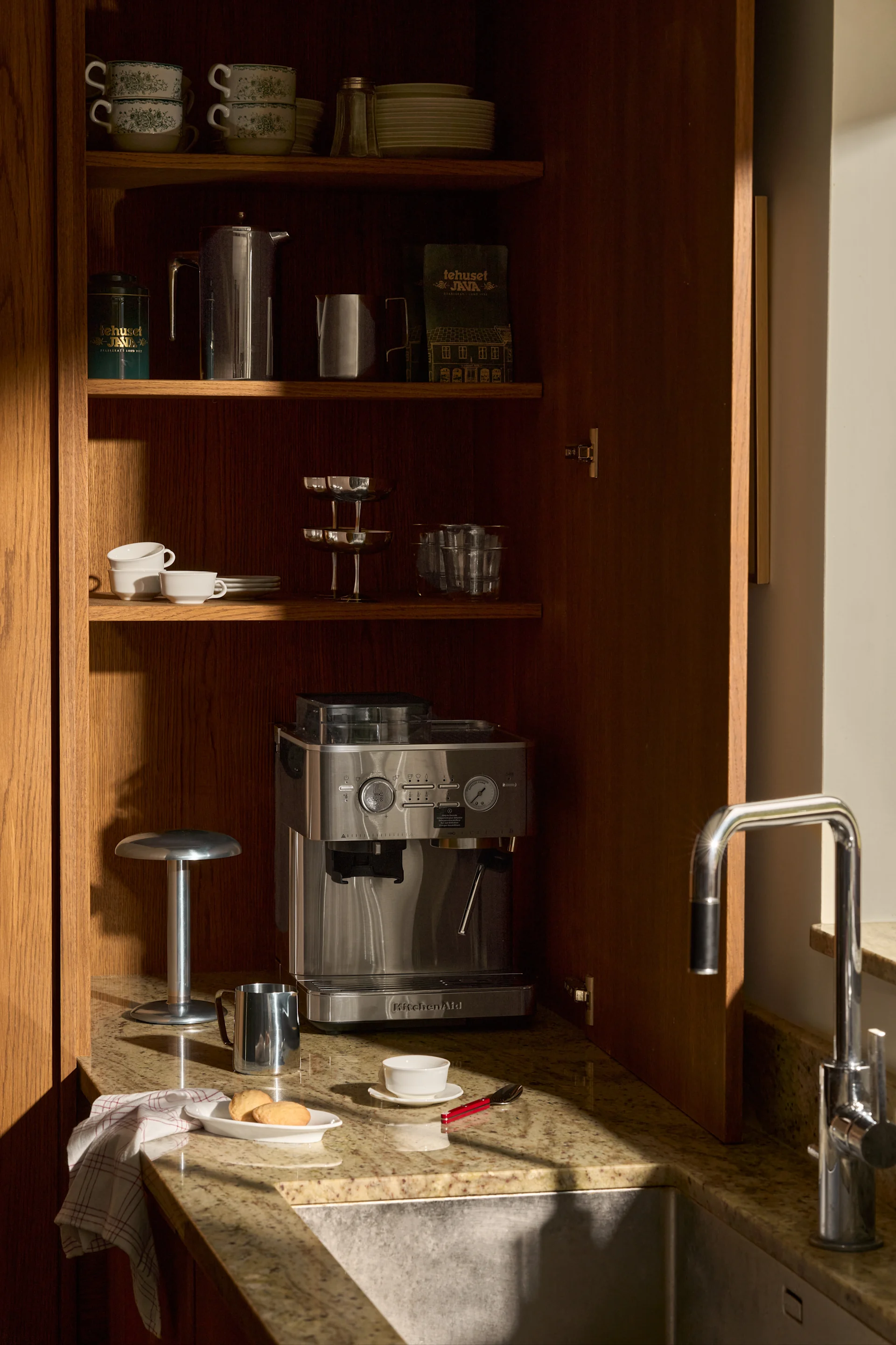 Avoin puukaappi: KitchenAid-espressokeitin, pinotut kupit, lautaset, kahvitarvikkeet ja allas graniittitasolla.