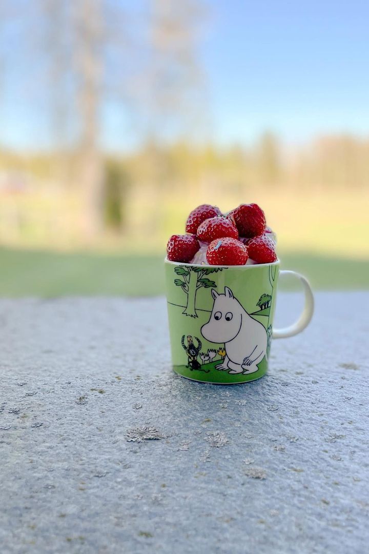 @mylittlemoominfamily:n vihreä Muumi-kuppi on ulkona lattialla, täynnä mansikoita.