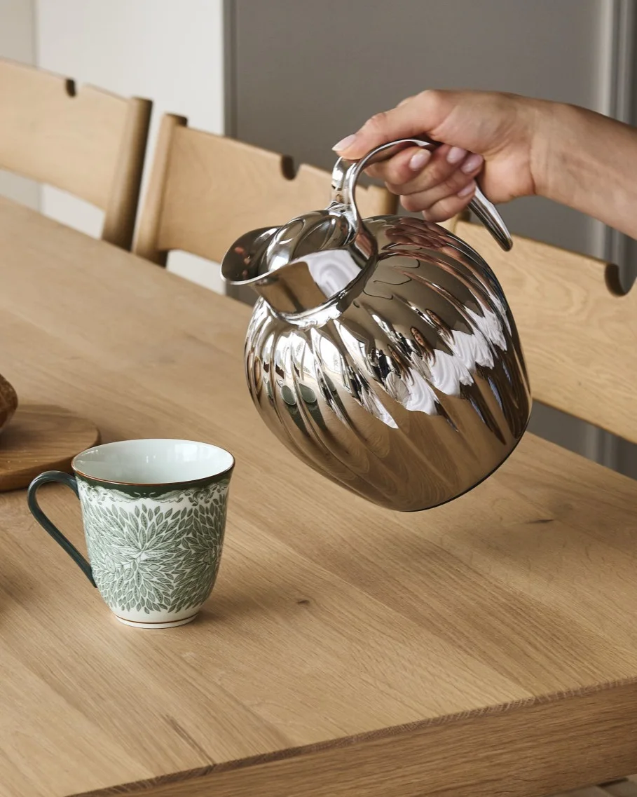 Käsi kaataa hopeisesta uurretusta karahvista valkoiseen mukiin, jossa on vihreä lehtikuvio, puupöydällä.