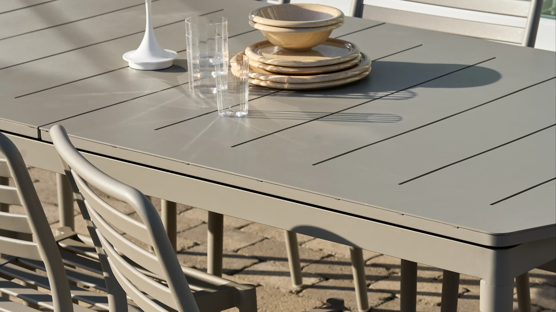 Vaaleanharmaa säleikköinen ulkoruokapöytä, jossa pinottuja beigenvärisiä lautasiasetteja ja kulhoja, kaksi kirkasta lasia ja valkoinen koriste-esine.