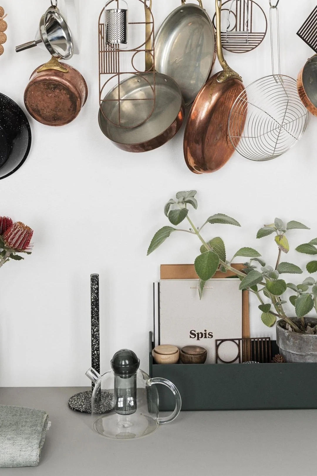 Ferm Livingin Still-teekannu on yksinkertainen mutta tyylikäs, ja se seisoo tässä keittiön työtasolla, ja sen yläpuolella roikkuu kattiloita.