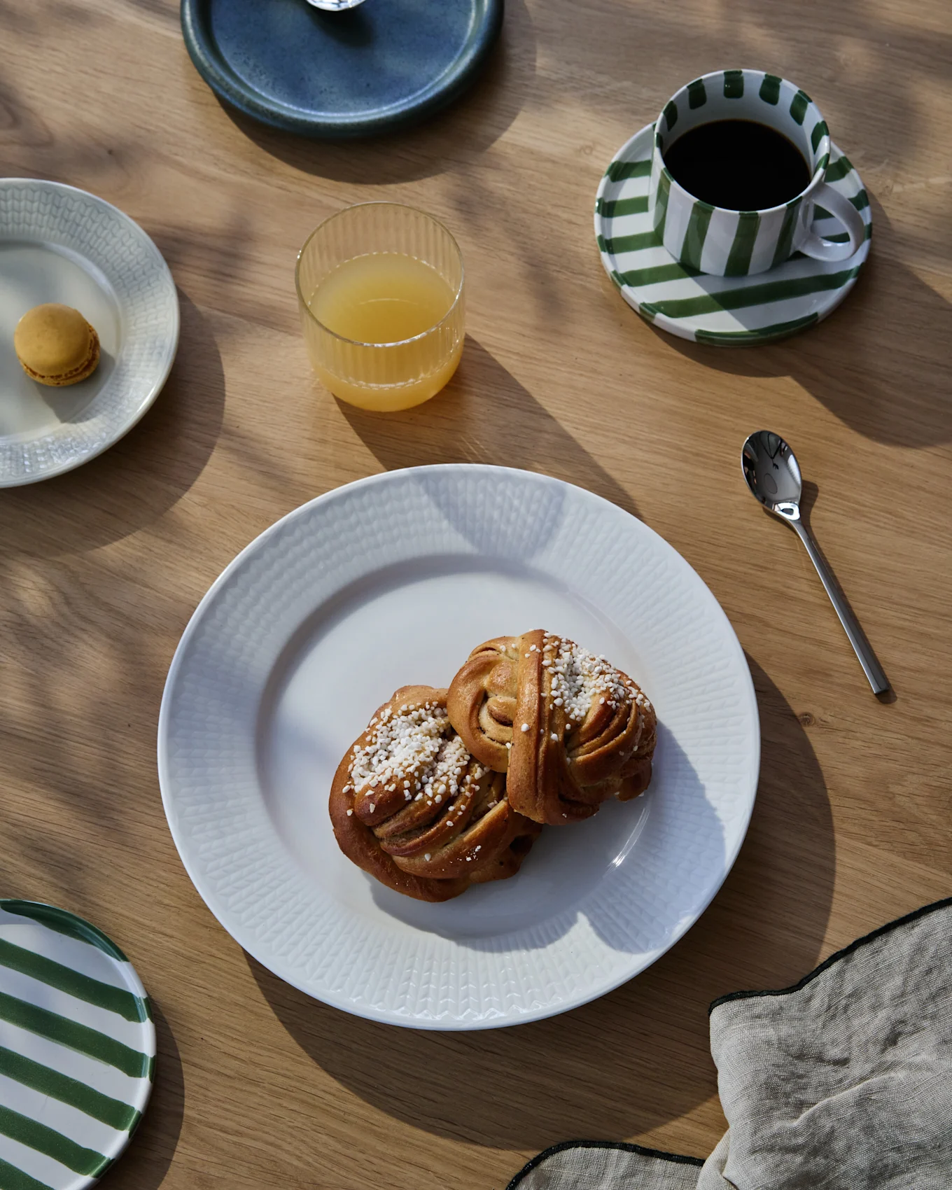 Puupöydällä aamiaiskattaus, jossa kaksi korvapuustia, kahvia raidallisessa kupissa, appelsiinimehua ja macaron-leivos.