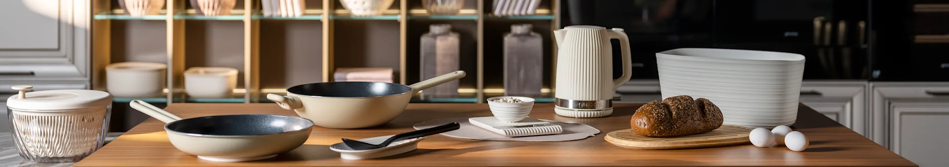 Keittiötaso, jolla on beigenvärisiä paistinpannuja, valkoinen uurrettu vedenkeitin, siemenleipä leikkuulaudalla ja munia. Moderneja keittiötarvikkeita.