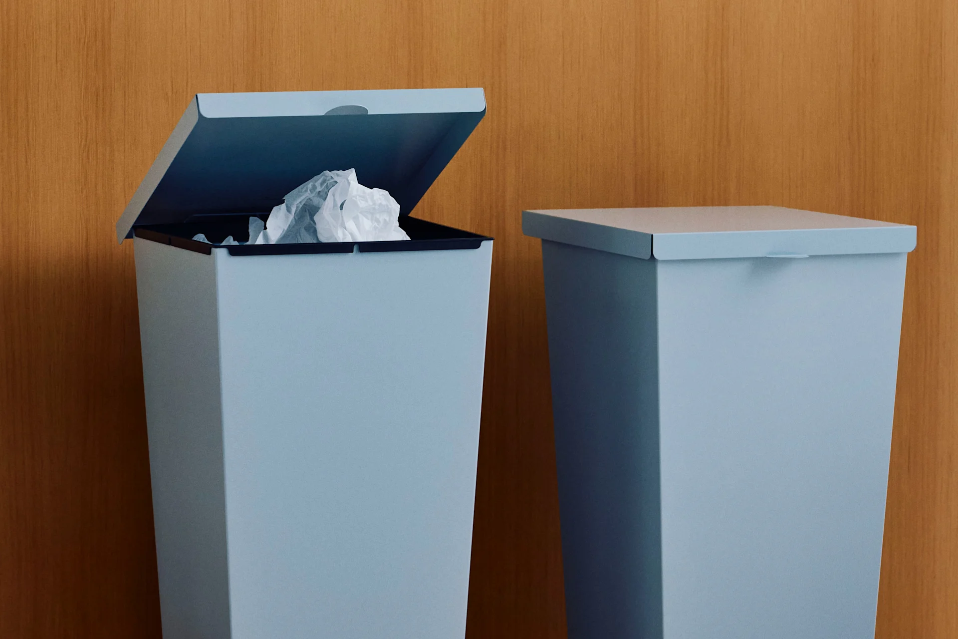 Kaksi vaaleansinistä roskakoria puuseinää vasten, toinen auki ja siinä rypistettyä paperia.