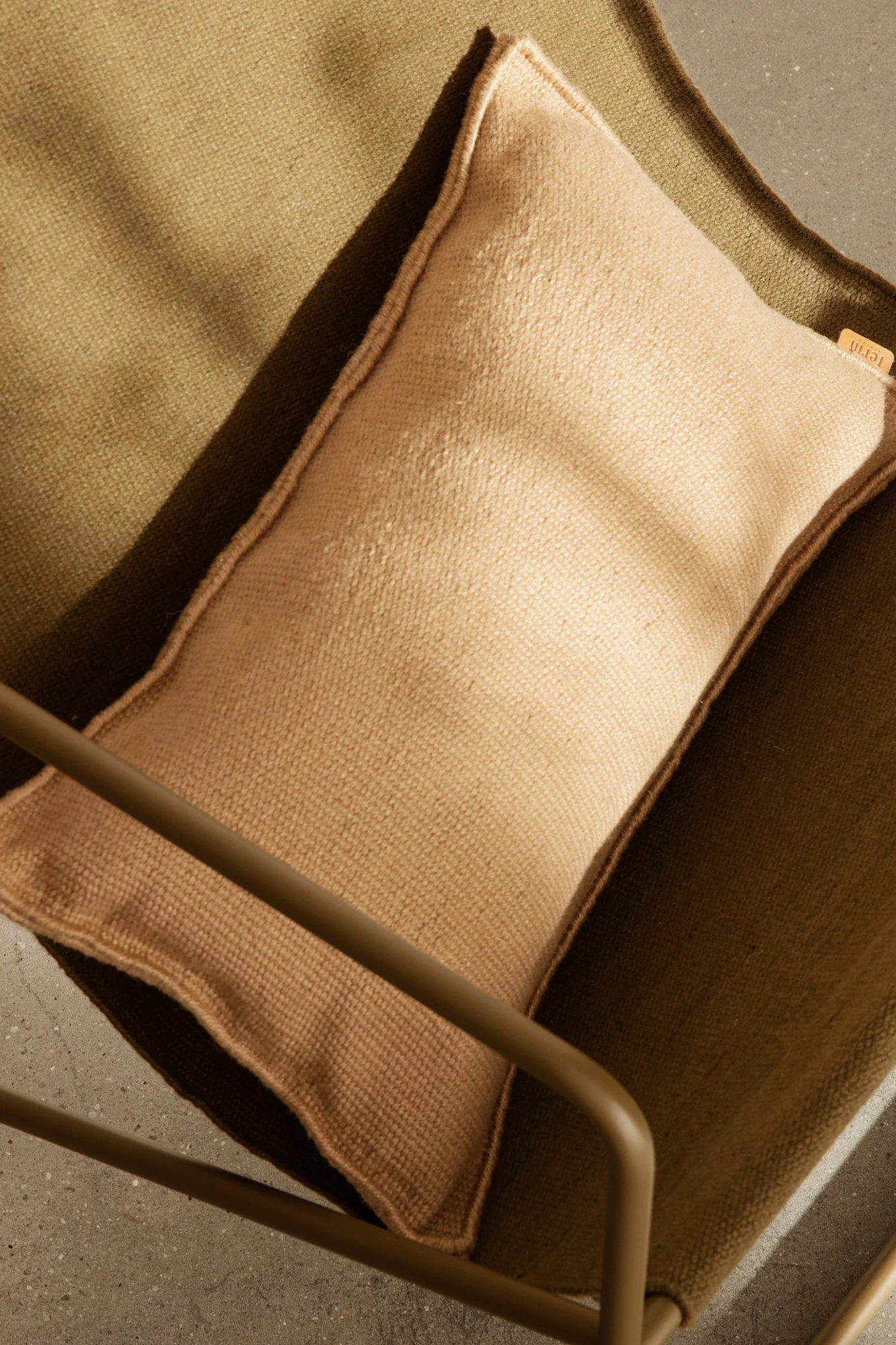 Ylhäältä kuvattu vaaleanruskea tyyny tumman oliivinvihreällä kangastuolilla, osa metallirunkoa ja betonilattia näkyvissä.