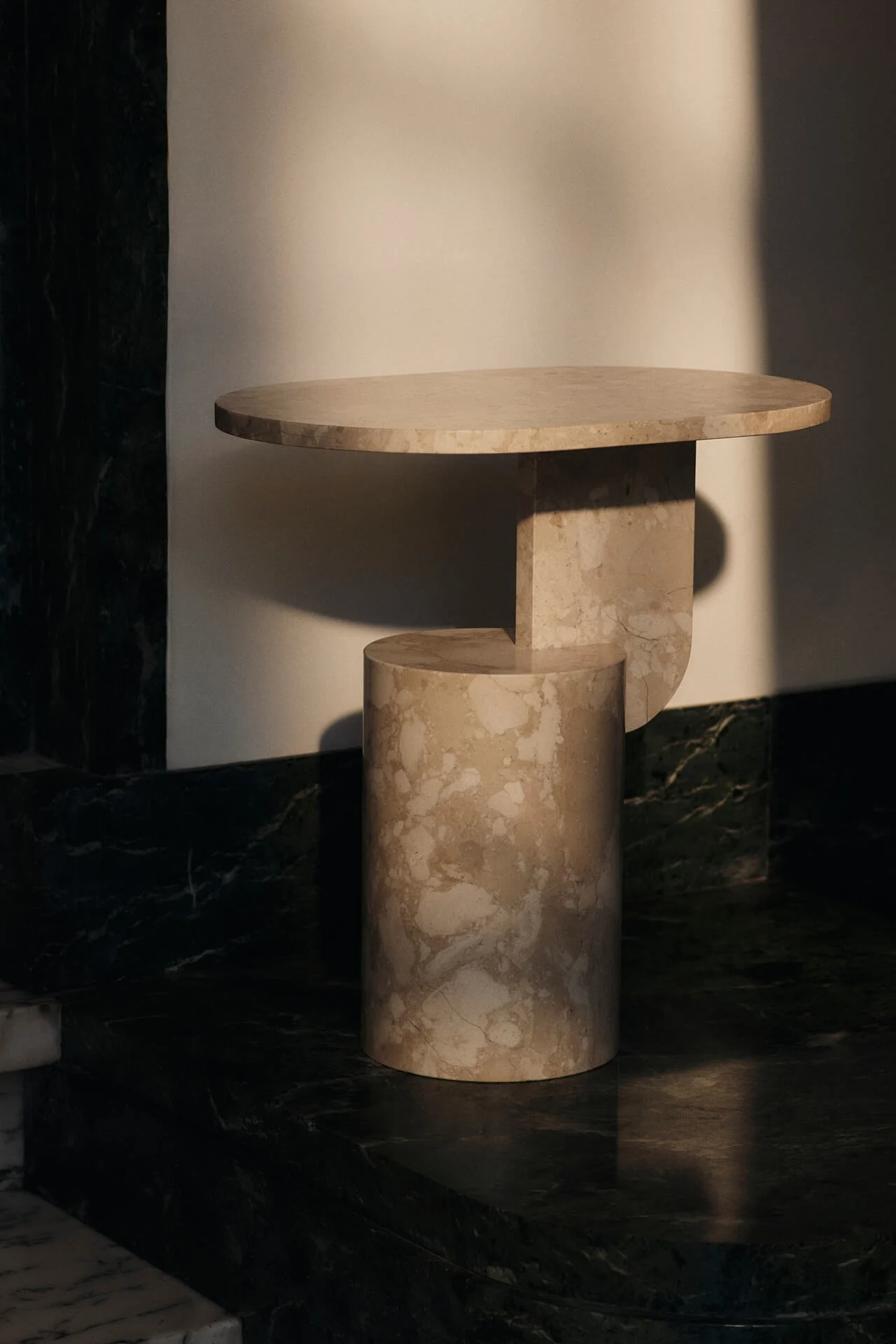 Moderni beige marmorinen sivupöytä, jossa pyöreä kansi ja geometrinen jalka vaaleaa seinää vasten.