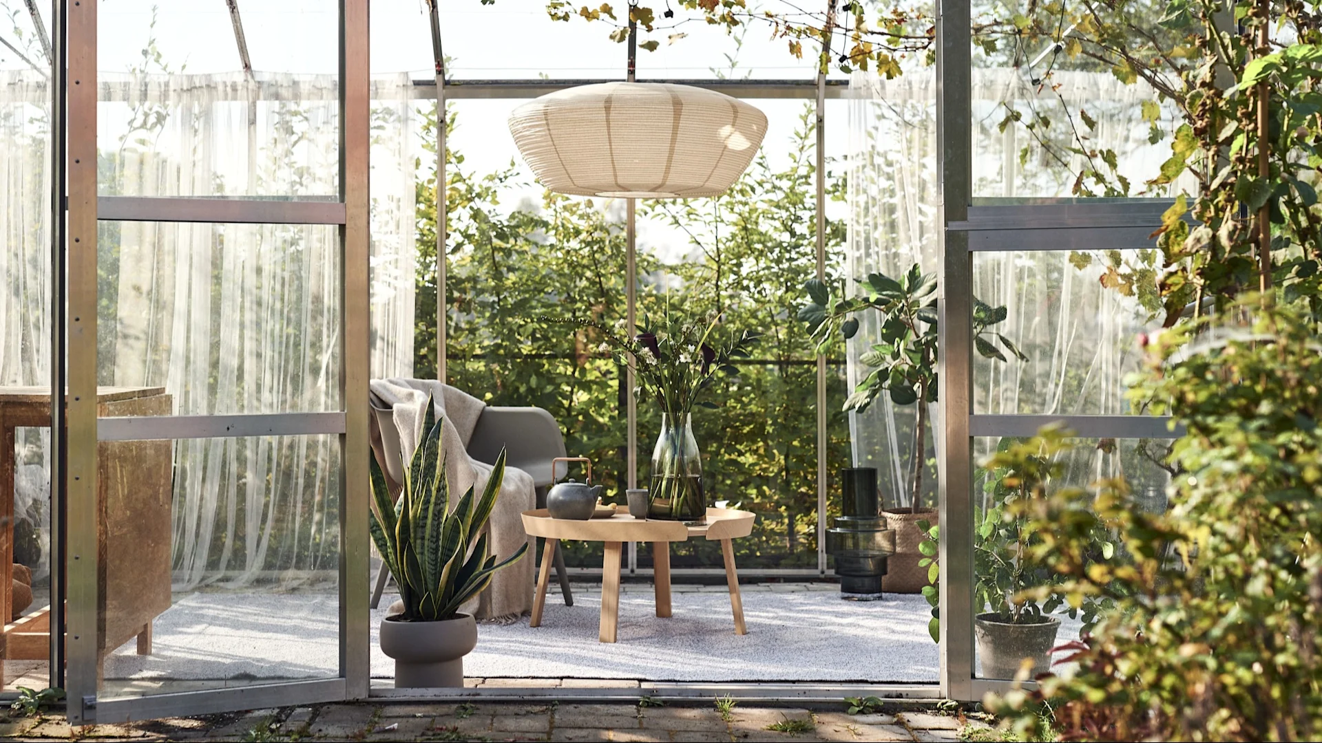 Ulkomatto: Tässä näet kasvihuoneen, joka on muutettu viihtyisäksi olohuoneeksi riippuvalaisimella, tuolilla, sivupöydällä ja suurella ulkomatolla.