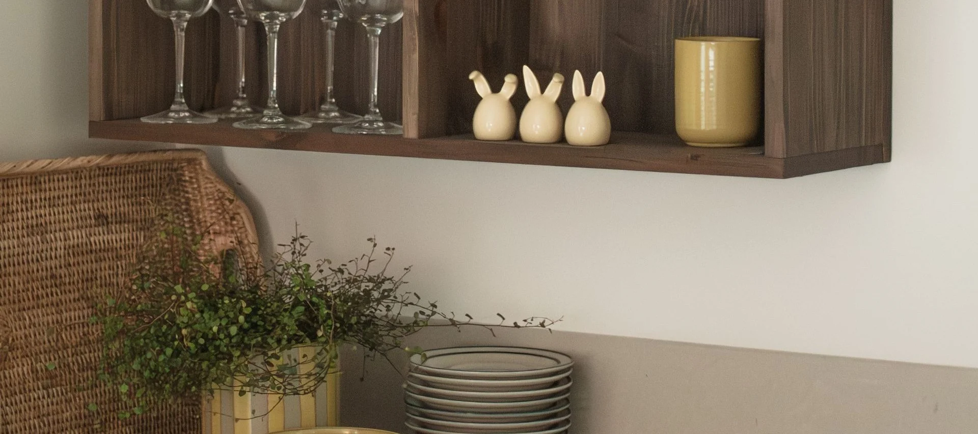 Puinen hyllykkö, jossa laseja, kolme pupuhahmoa ja keltainen muki, alla pajuinen kori ja kasveja.