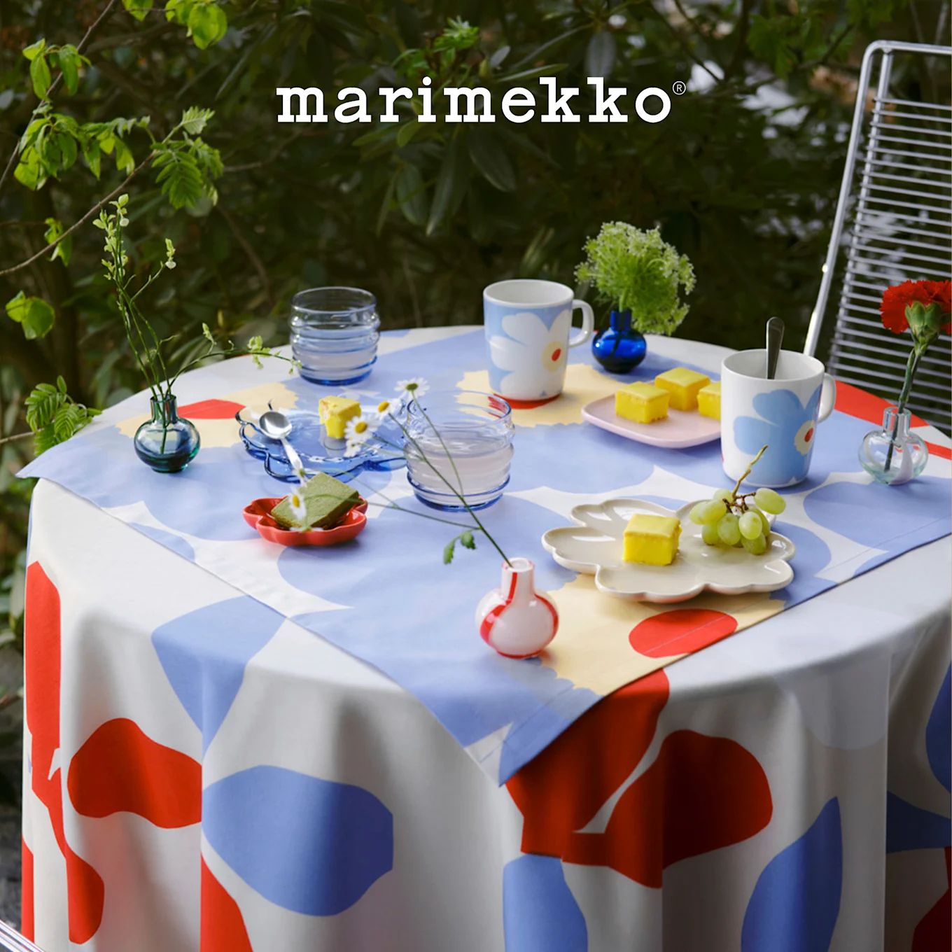 Ulkopöytä Marimekko punaisella, sinisellä, valkoisella kuvioidulla pöytäliinalla, katettuna kukkamukeilla, jälkiruoilla ja pienillä kasveilla. Marimekko-logo.