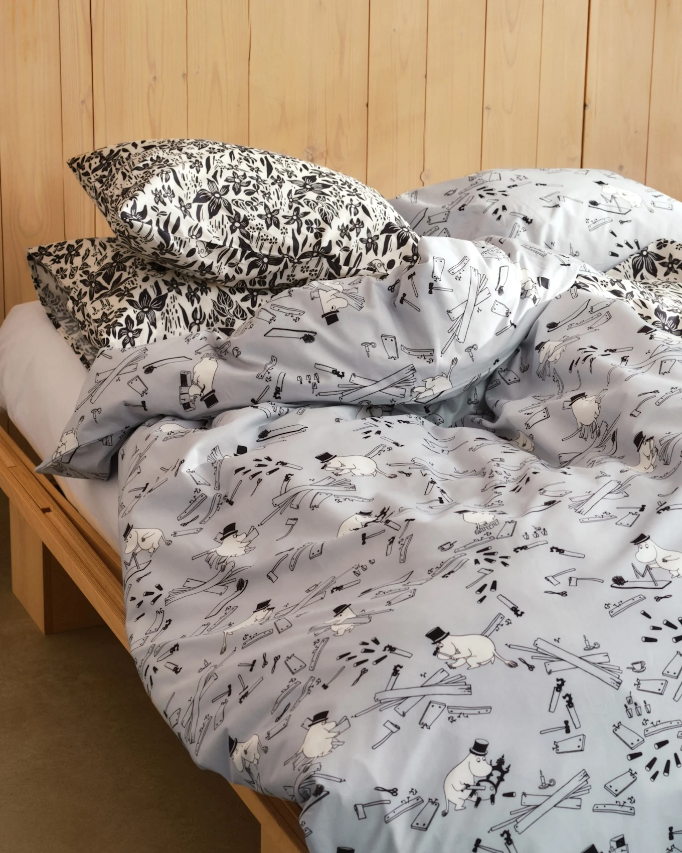 Sänky, jossa mustavalkoiset kukkakuvioiset tyynyt ja vaaleansininen pussilakana sarjakuvahahmoilla, työkaluilla ja puulankuilla. Puinen sängynrunko.