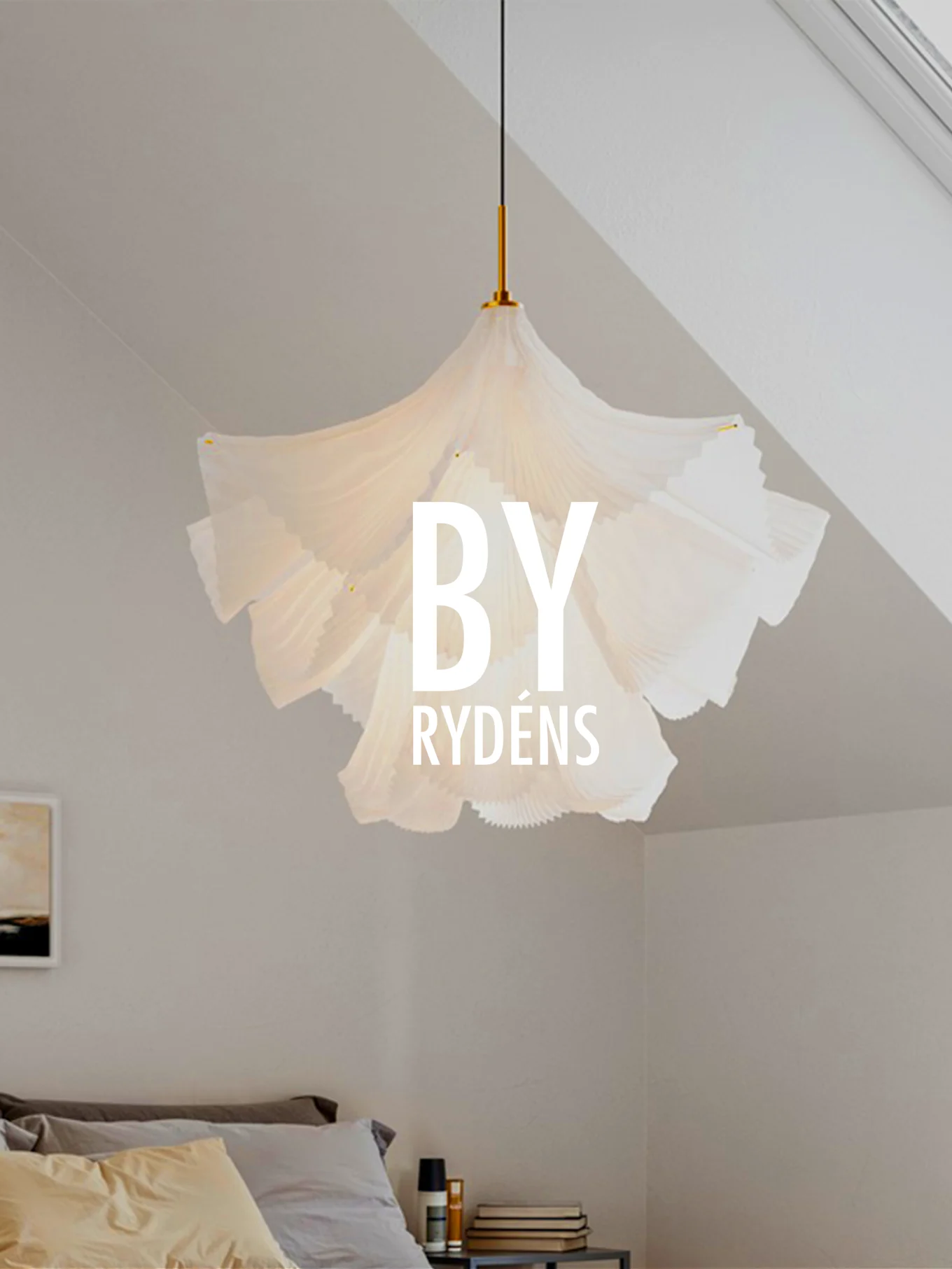 Valkoinen, veistoksellinen laskostettu riippuvalaisin roikkuu sängyn yllä tyynyjen ja yöpöydän kanssa. Teksti 'BY RYDÉNS' on valaisimen päällä.