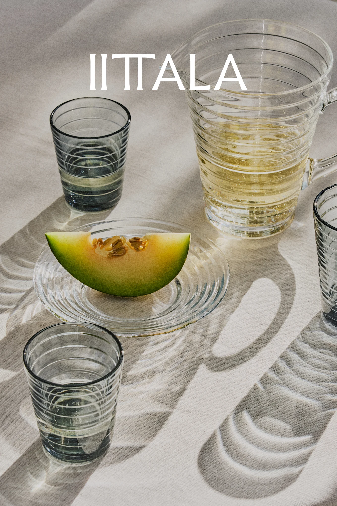 IITTALA-logo lasien, kuohuvan juoman kannun ja meloninpalan päällä lautasella, vaalealla kankaalla.