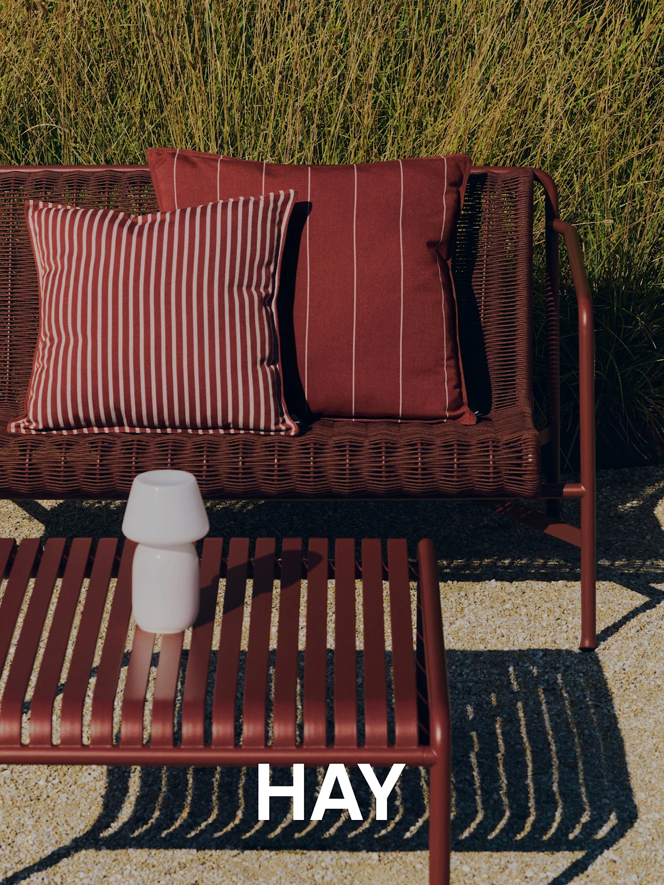 Ulkoilmanäkymä, jossa punainen punottu sohva, raidalliset tyynyt, säletty sohvapöytä valkoisella lampulla ja korkeaa ruohoa taustalla. HAY-logo.