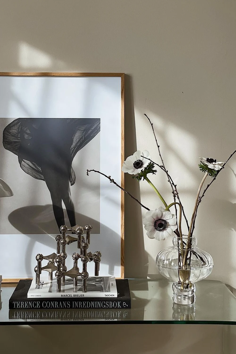 Kehystetty mustavalkoinen valokuva jaloista ja hulmuavasta kankaasta, hopeinen modulaarinen kynttilänjalka, kirjoja sekä valkoisia vuokkoja lasimaljakossa.