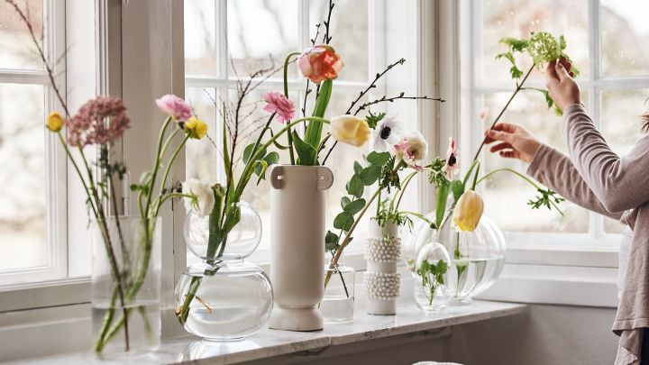 Ikkunalaudalla on kokoelma kirkaslasisia maljakoita, joissa on värikkäitä kevätkukkia. 