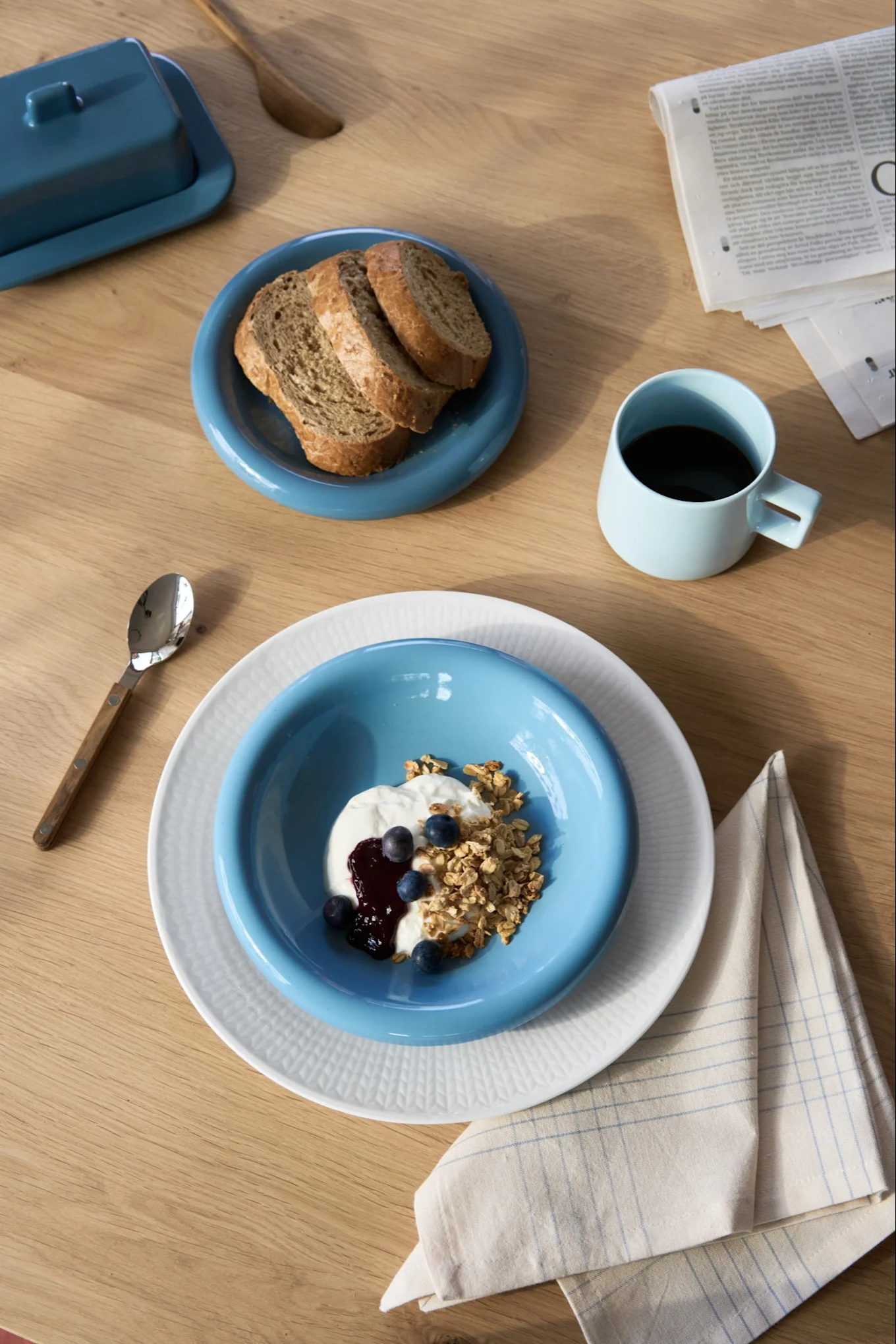 Aamiainen: jogurtti, granola, mustikat, leipä sinisillä lautasilla, kahvia sinisessä mukissa, voikuppi puupöydällä.