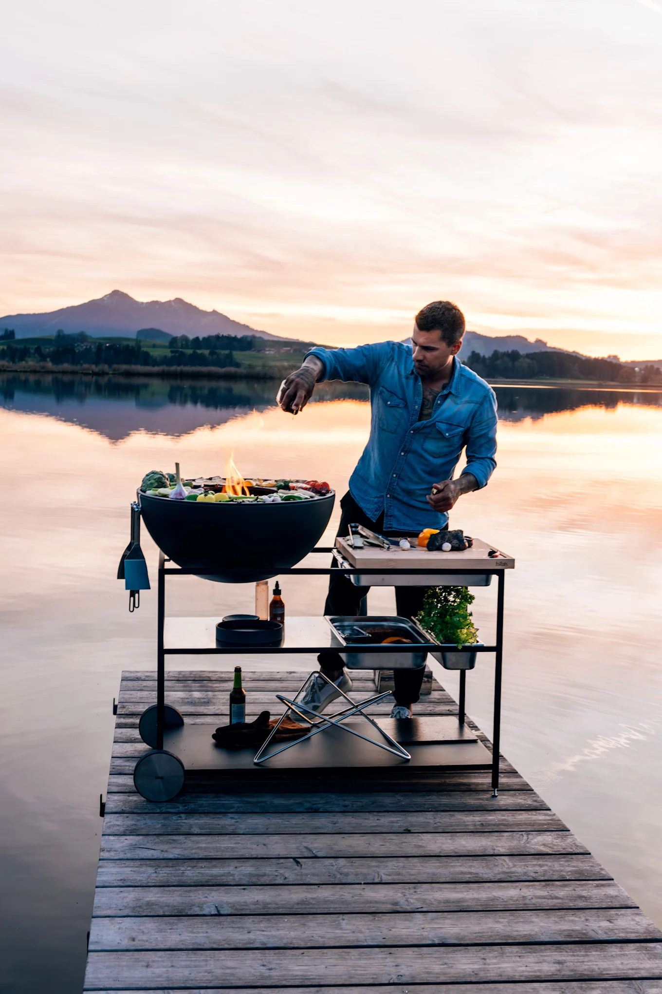 Mies farkkupaidassa grillaamassa ruokaa pyöreällä mustalla grillillä ja valmistelupöydällä puulaiturilla auringonlaskun aikaan.