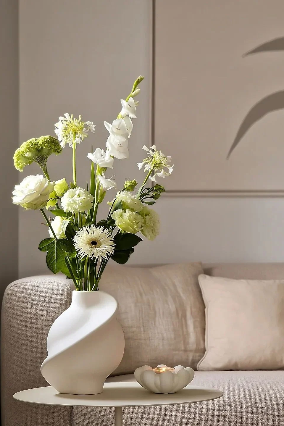 Beigenvärisessä olohuoneessa on sivupöytä, jota koristaa maljakko leikkikukilla ja teelyhty.