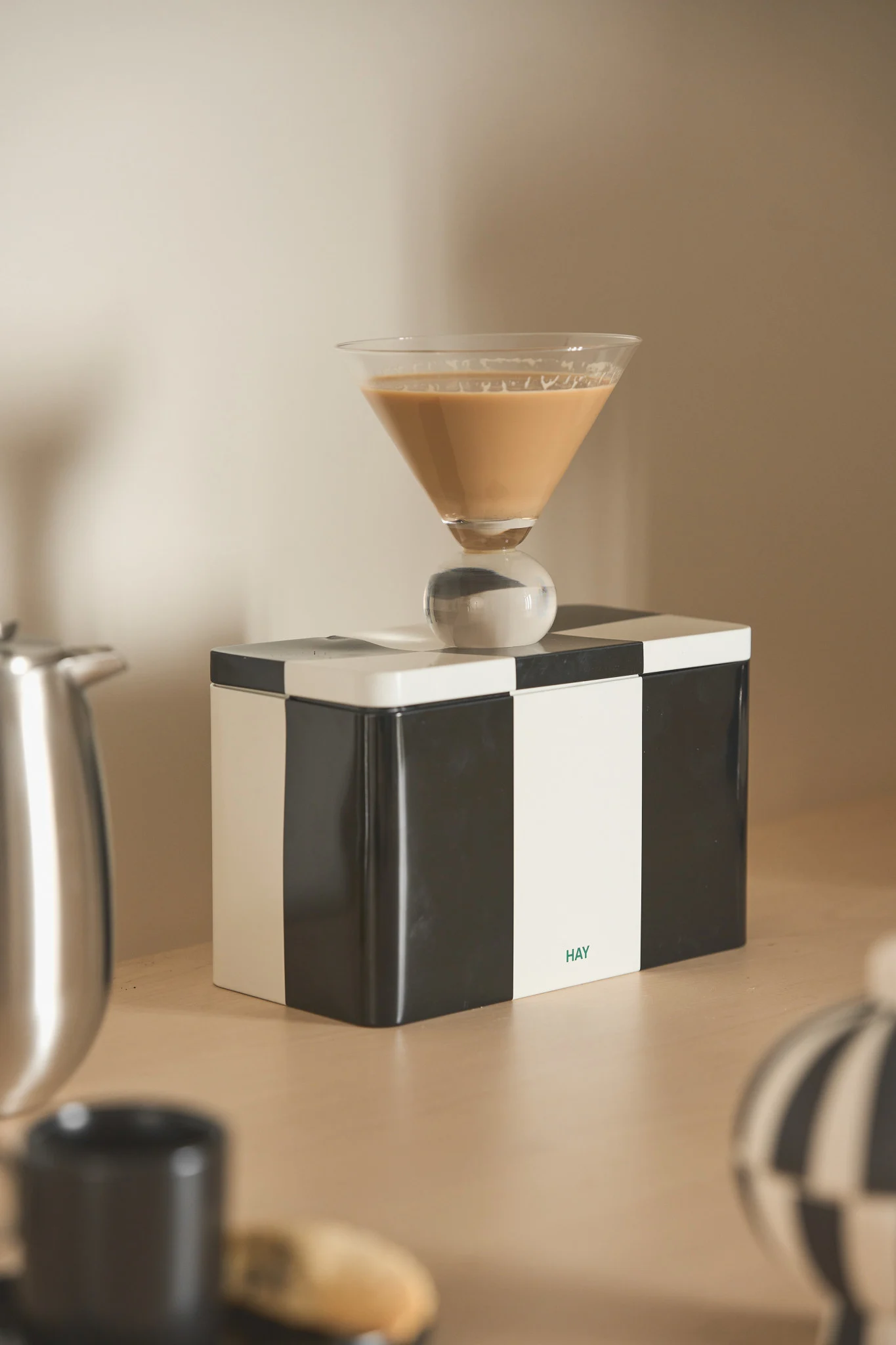 Martinilasi vaaleanruskealla juomalla ja pallomaisella jalalla mustavalkoisen ruudullisen laatikon päällä, jossa on 'HAY'-logo.