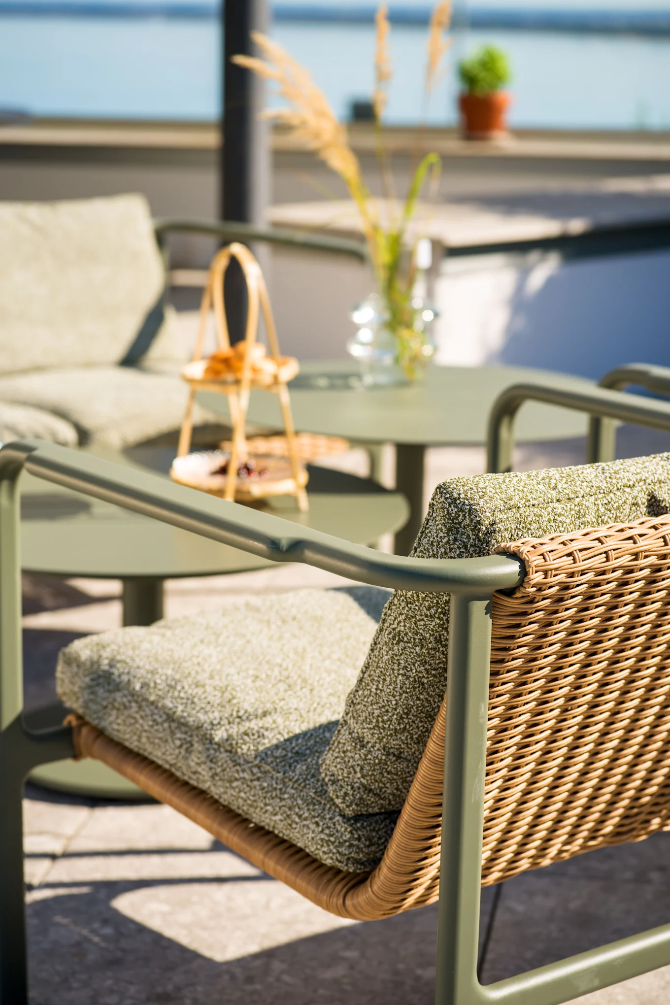 Ulkoterassin tuoli oliivinvihreällä rungolla, punotulla rottinkiselkänojalla ja vihreillä tyynyillä. Taustalla veden äärellä näkyy epätarkasti yhteensopiva pöytä ja tuoli.