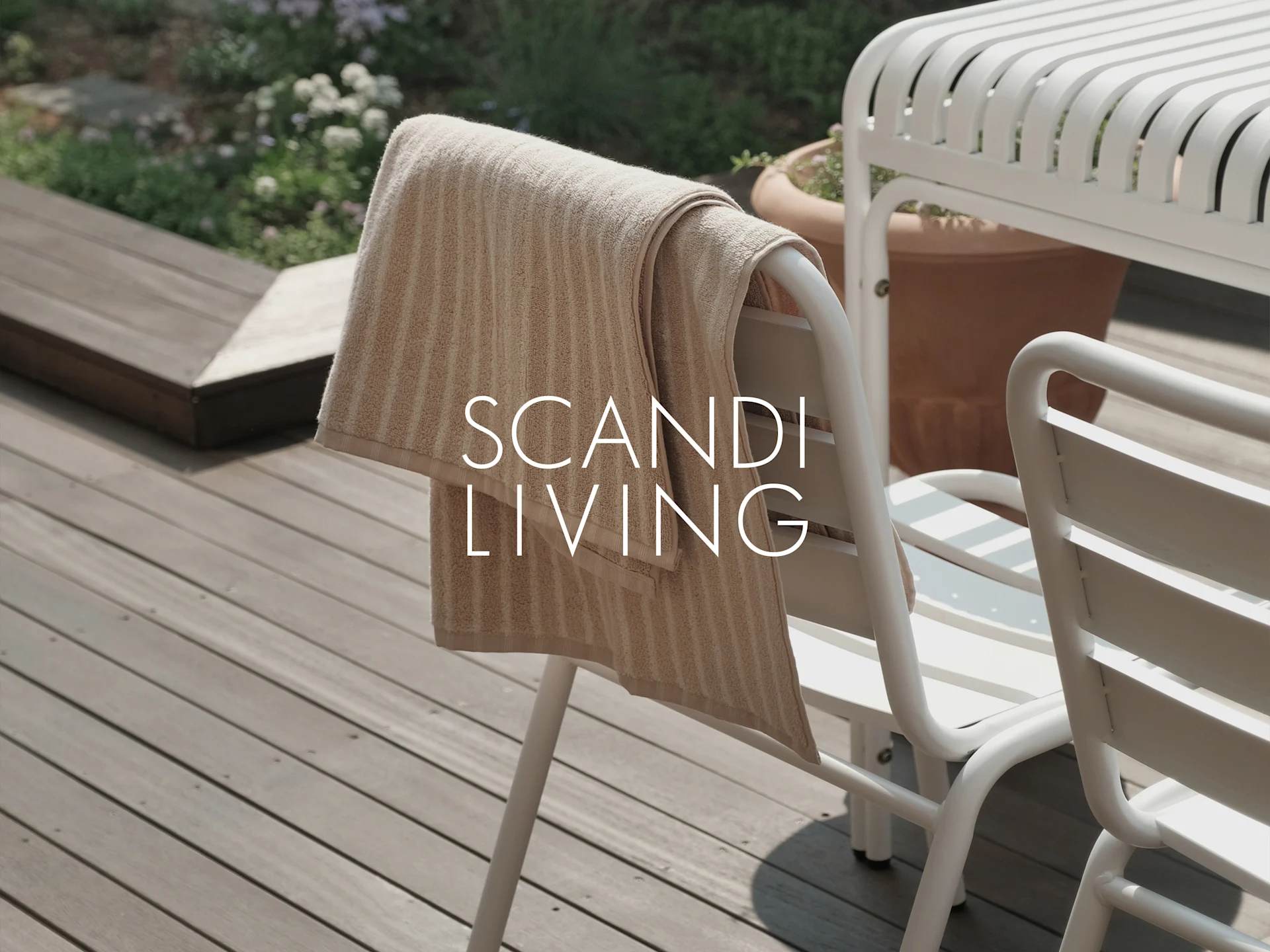 Beige raidallinen pyyhe roikkuu valkoisessa ulkotuolissa puuterassilla kasvien kanssa. Teksti: SCANDI LIVING.