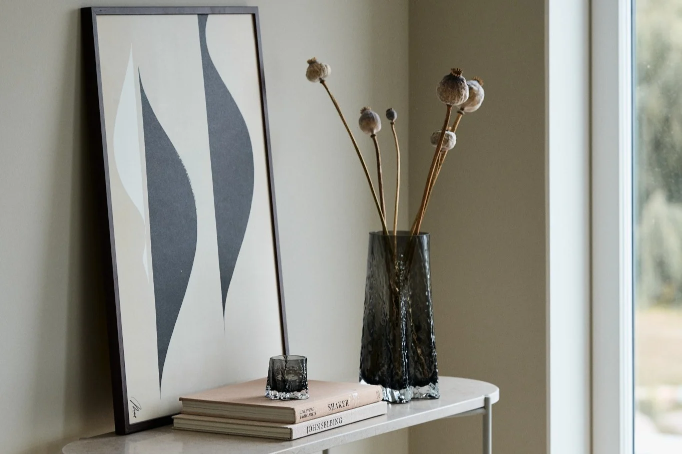 Tässä näet 30-senttisen Gry-maljakon ja Gry-kynttilälyhdyn, molemmat Cooee Designin värissä Smoke, seisomassa sivupöydällä julisteen edessä.  