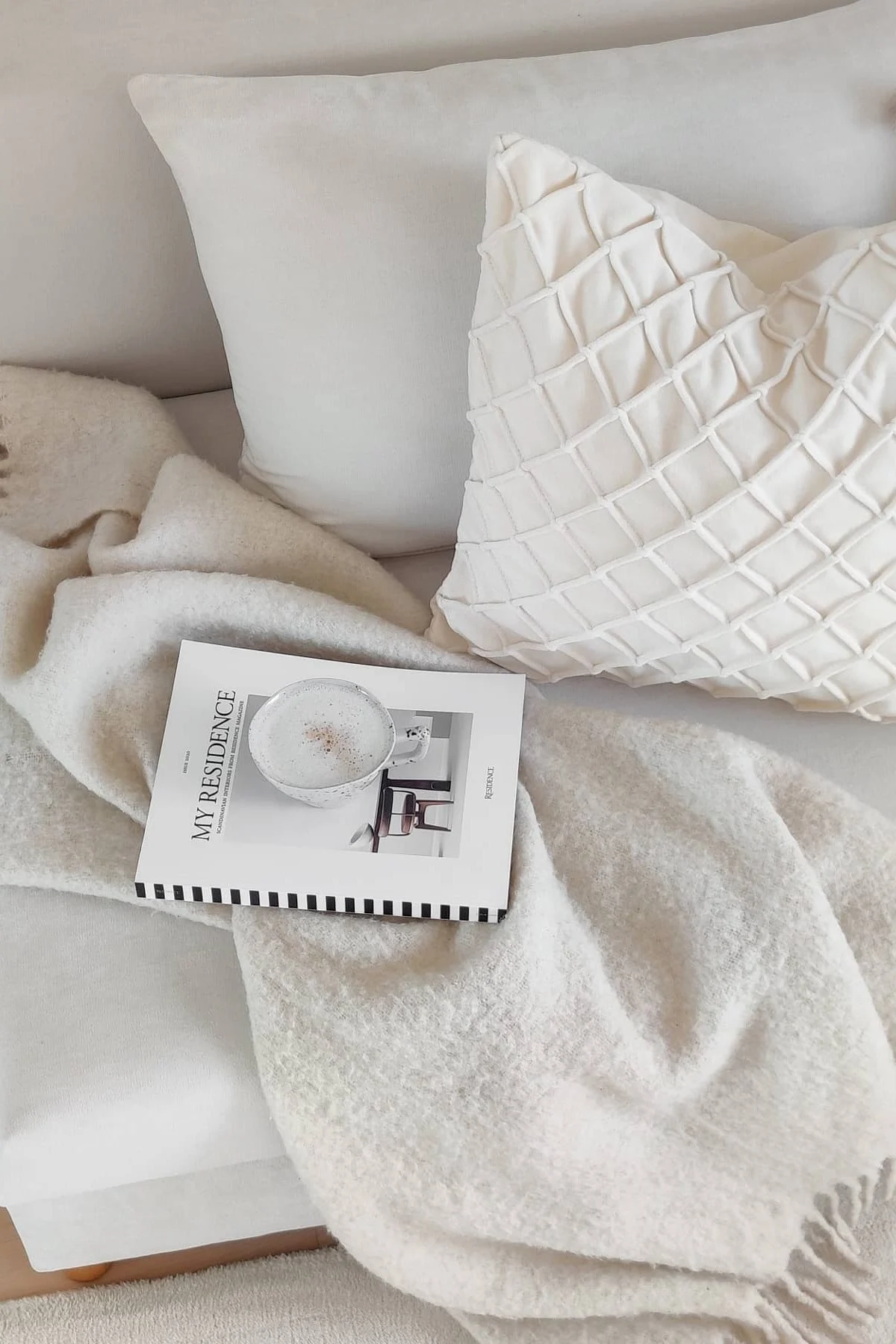 Ystävänpäivälahjat: Valkoisella sohvalla on kaksi valkoista tyynyä Chhatwal & Jonssonilta, Ralph-peitto Klippan Yllefabrikilta, kirja ja kuppi kahvia.