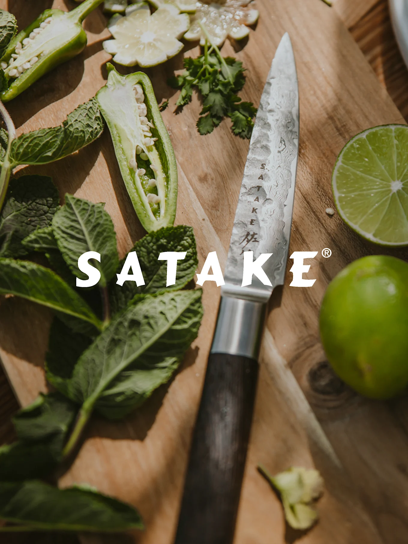 Satake-veitsi kuvioidulla terällä ja tummalla kahvalla leikkuulaudalla, ympärillään tuoretta chiliä, minttua ja limeä.