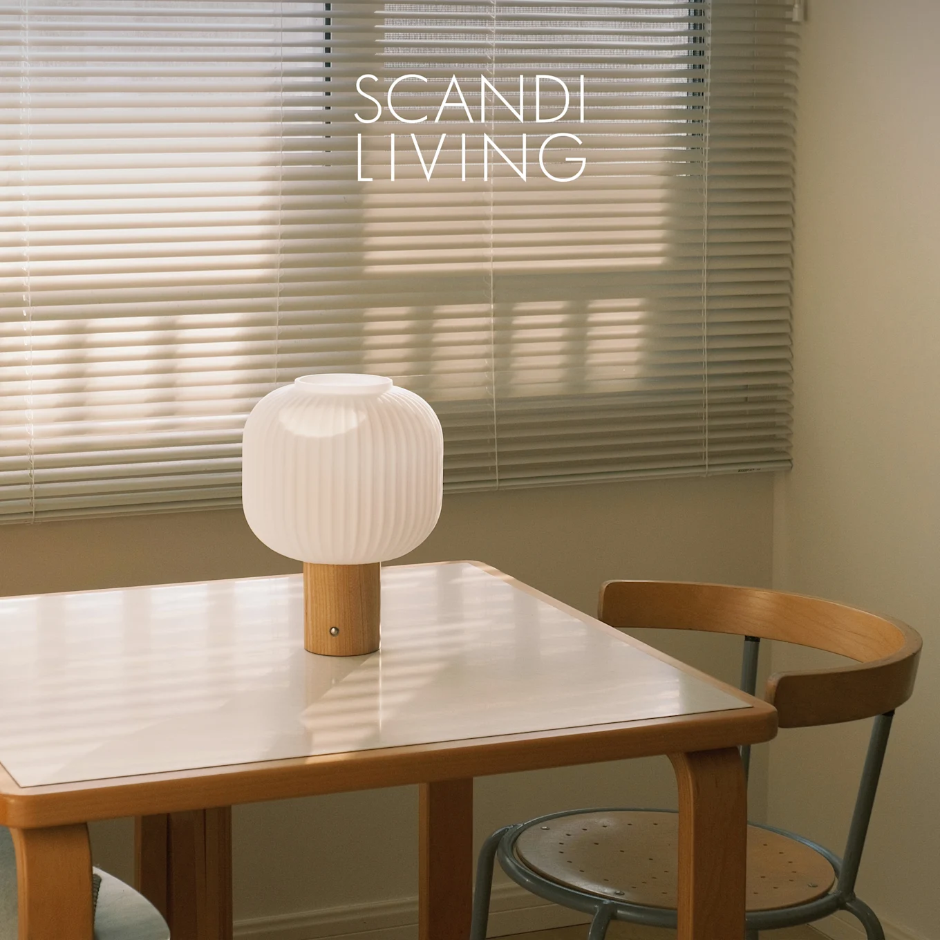 Valkoinen uritettu pöytävalaisin puujalalla valkoisella pöydällä ja tuolilla, ikkunakaihtimien edessä. Teksti: SCANDI LIVING.