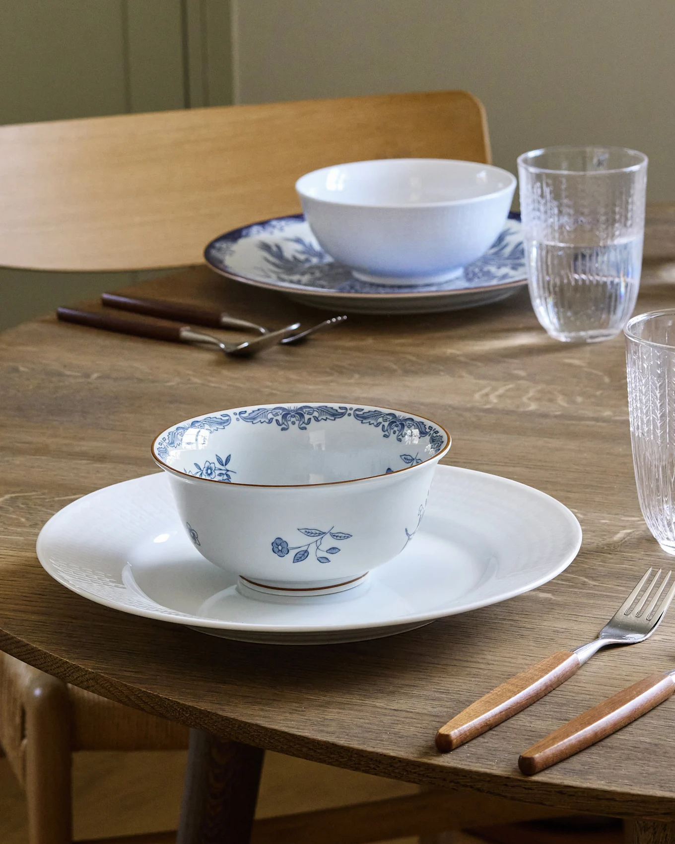 Ruokapöytä kahdelle katettuna valkoisilla ja sinikuvioisilla kulhoilla valkoisten lautasten päällä, puukahvaisilla aterimilla ja vesilaseilla.