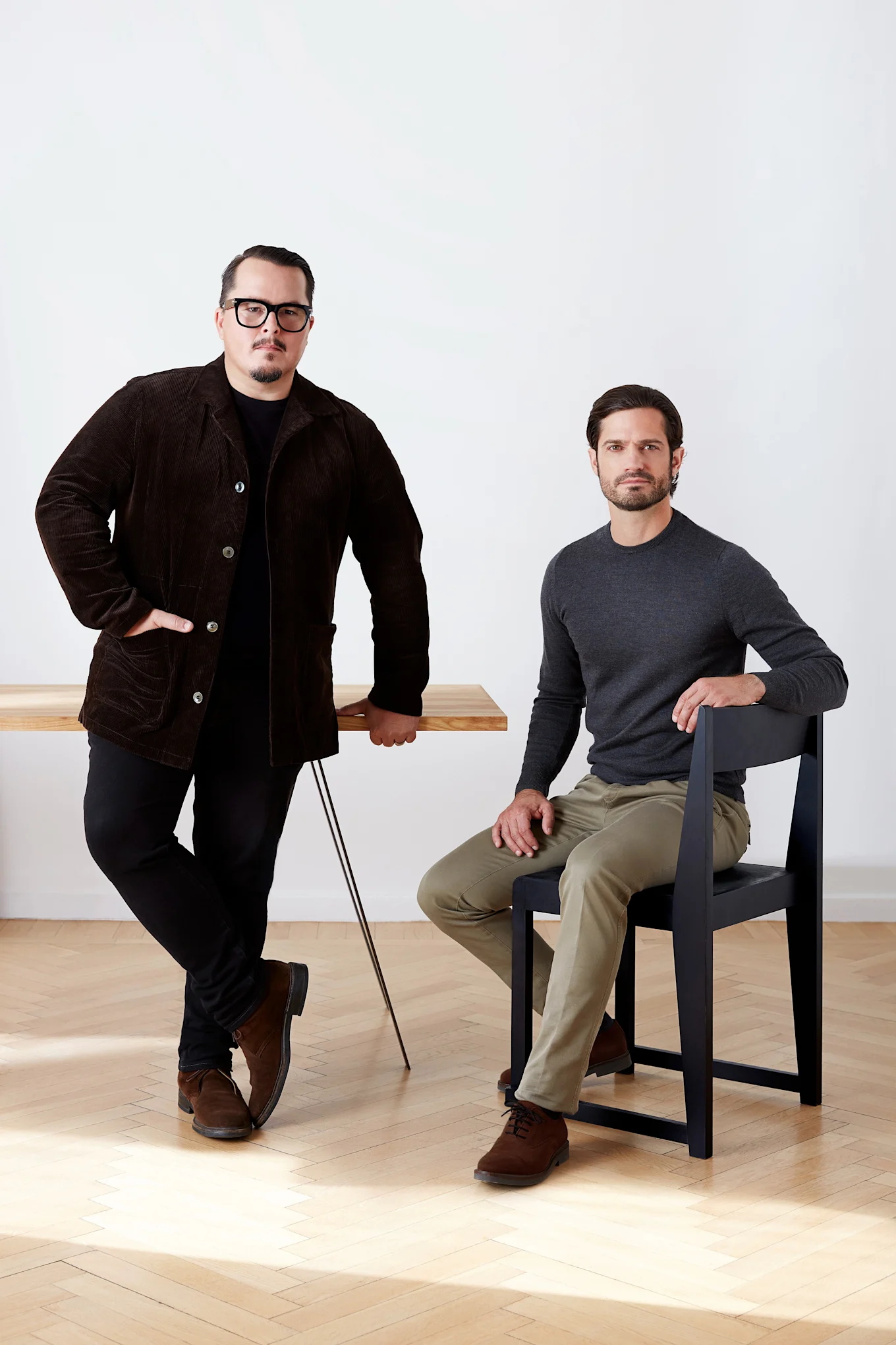 Kaksi miestä, toinen seisoo puupöydän ääressä ruskeassa vakosamettitakissa, toinen istuu mustalla tuolilla harmaassa paidassa.