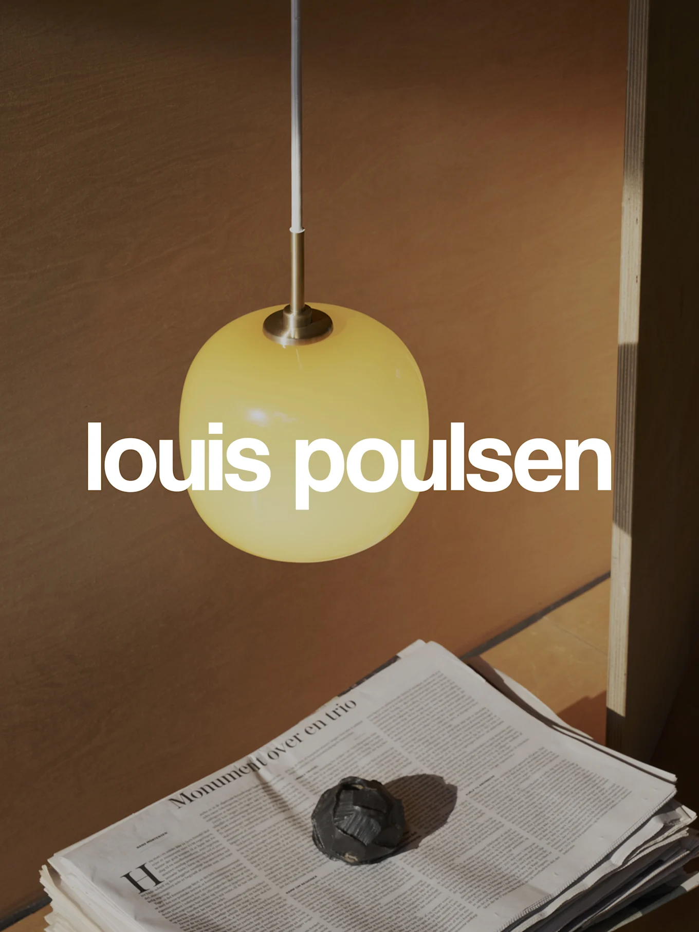Louis Poulsen -logo lämpimässä kohtauksessa, jossa keltainen pallomainen riippuvalaisin, pinottuja sanomalehtiä ja tumma koriste-esine.