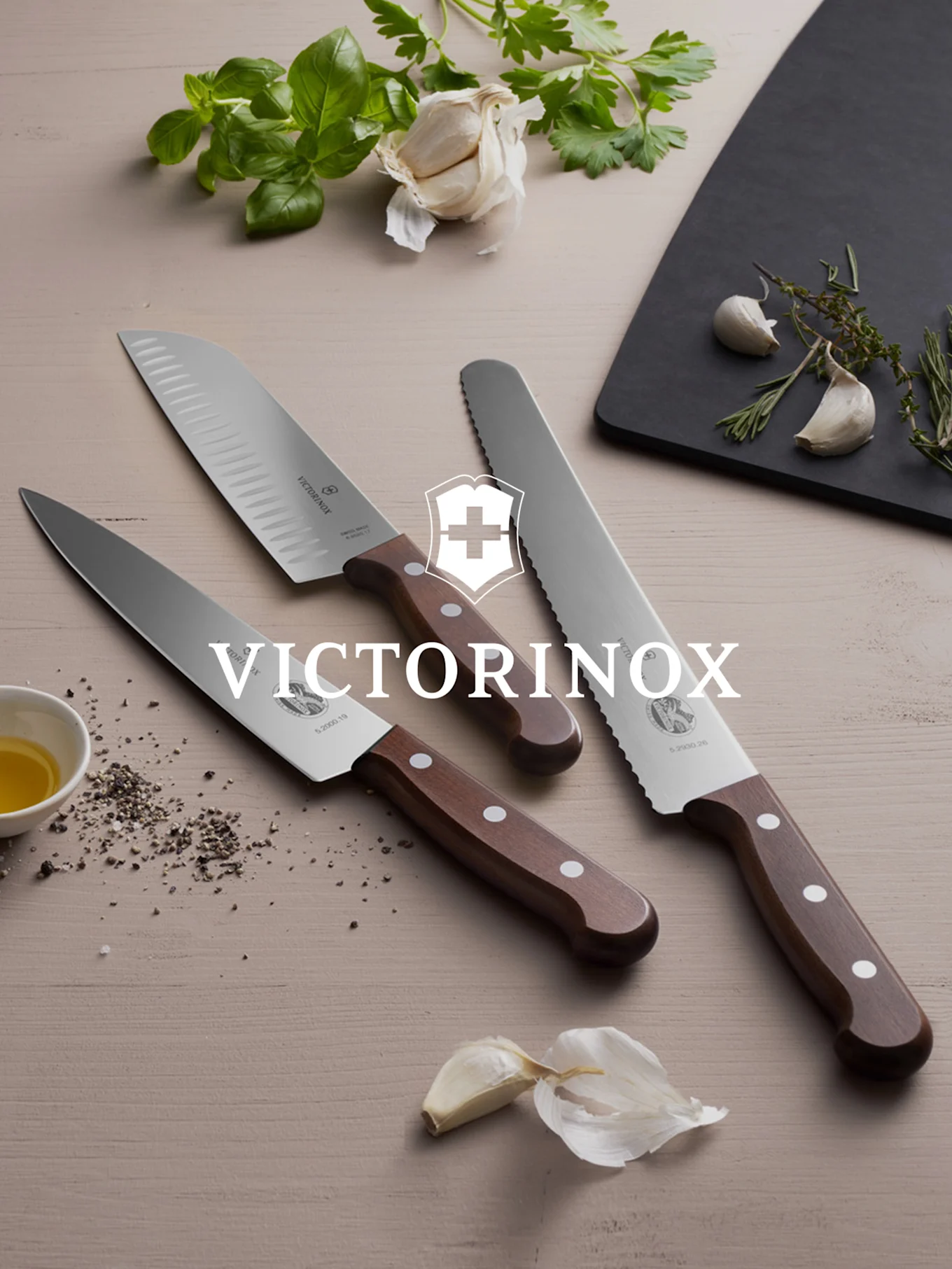 Kolme Victorinox-veistä puukahvoilla, tuoreita yrttejä ja valkosipulia puupinnalla.