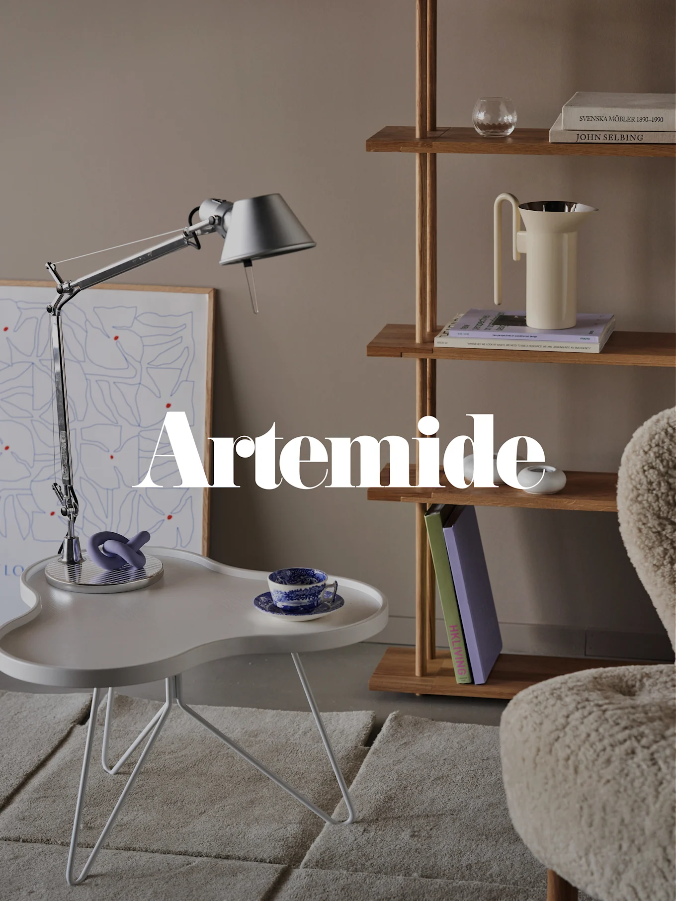 Moderni olohuone, jossa hopeinen pöytälamppu, valkoinen sivupöytä, puinen kirjahylly ja kermansävyinen nojatuoli. Tekstipeite: 'Artemide'.