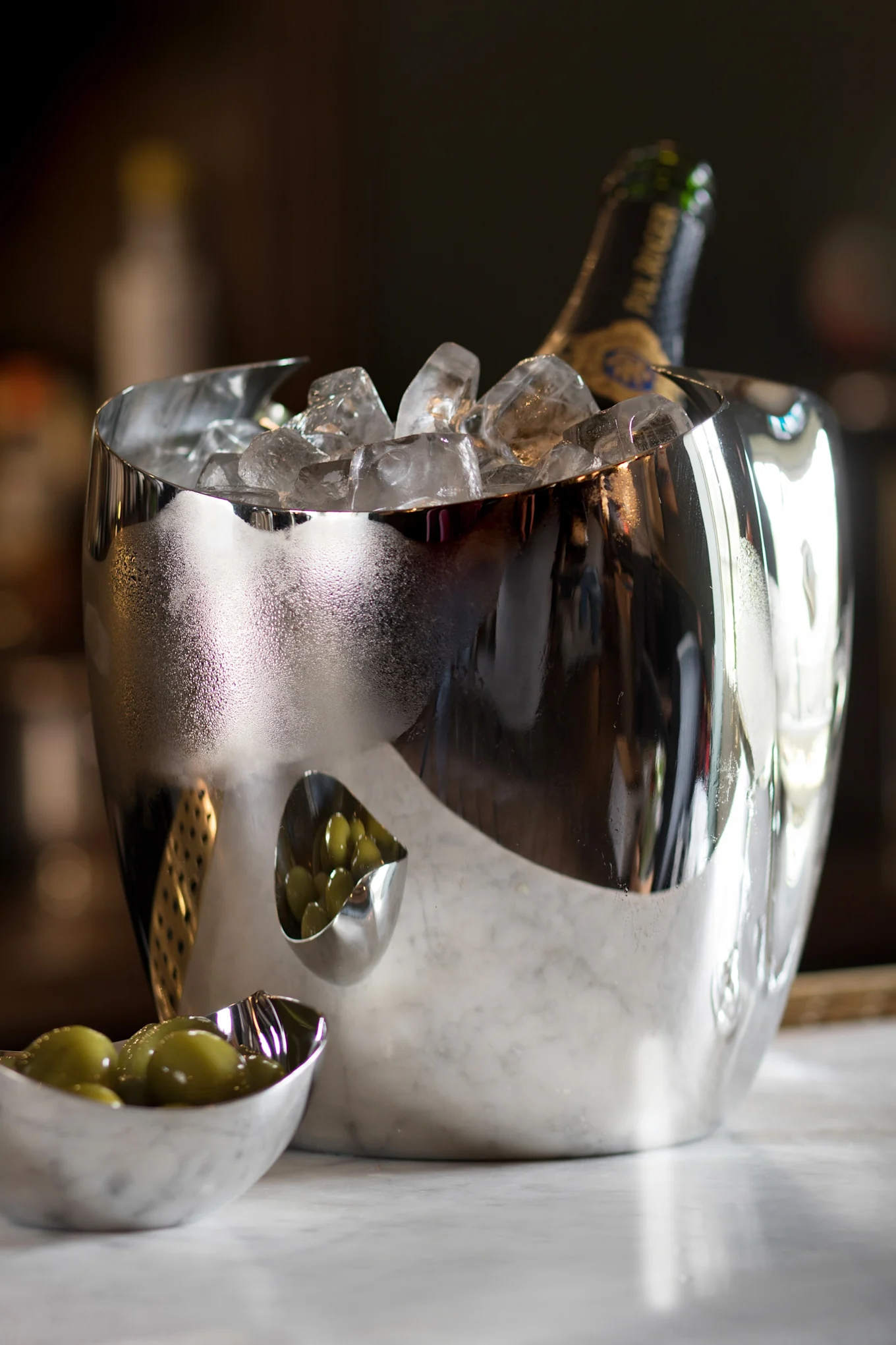 Heijastava hopeinen jääastia samppanjapullolla ja jäällä, kahden pienen vihreillä oliiveilla täytetyn kulhon vieressä marmorilla.