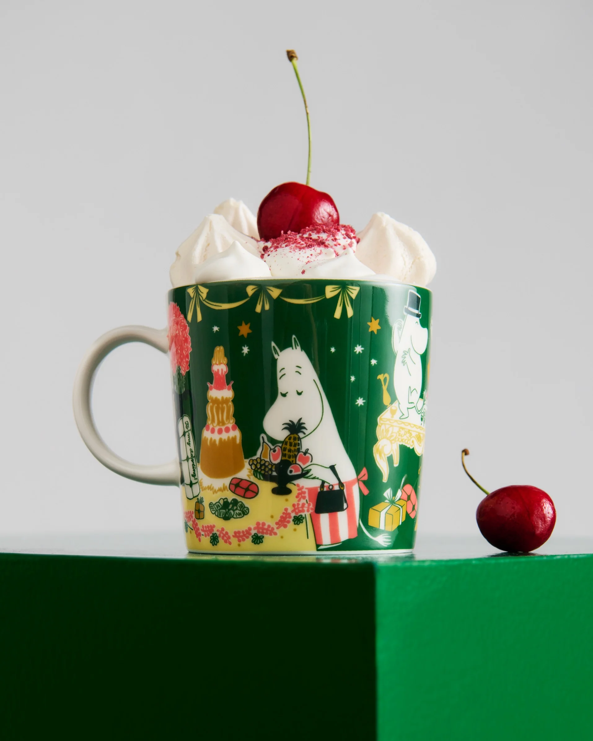 Tummanvihreä Moomin-muki, jossa kermavaahtoa, punaisia nonparelleja ja kirsikka. Toinen kirsikka lepää vihreällä alustalla.